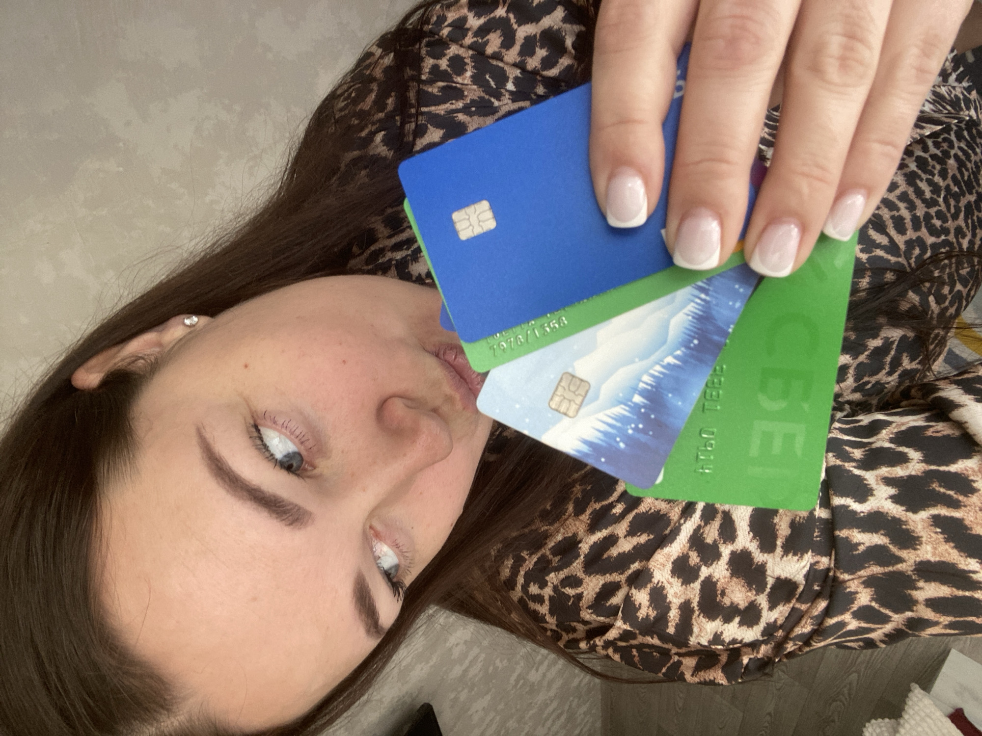A woman is holding three credit cards in her hand, positioned near her face. The top card is solid blue, the middle card features a winter mountain landscape, and the bottom card is green with some lettering visible. The woman has light skin, brown hair, and light-colored eyes. She is wearing a leopard-print top. The background appears to be a simple wall or neutral surface. The specific location is not identifiable from the image.