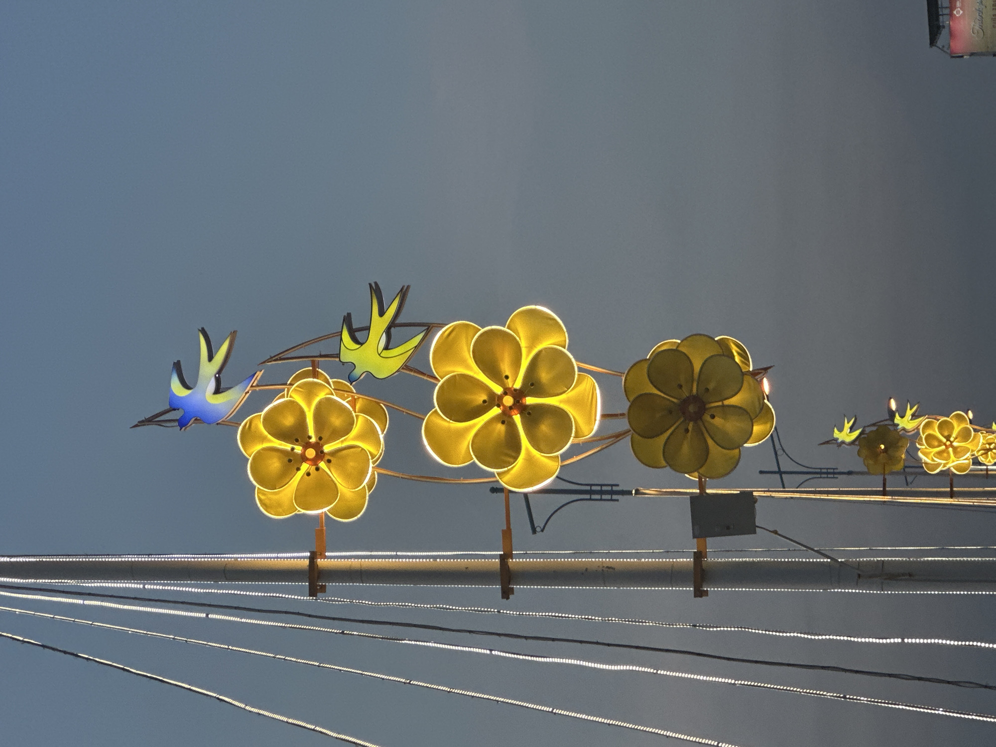 An upward-angle, medium shot captures a tall, metallic pole on the left, adorned with vertical strings of white LED lights. To the right of the pole, a series of illuminated yellow, five-petaled flower decorations hang vertically, attached by thin, brown wires. The largest flower is positioned at the top, with progressively smaller ones descending the pole. Interspersed between the flowers are stylized, light-yellow bird decorations with blue accents, appearing to fly. In the background, the sky is a muted, twilight blue. The scene is set against the backdrop of Nam Nha Trang Ward, Vietnam.