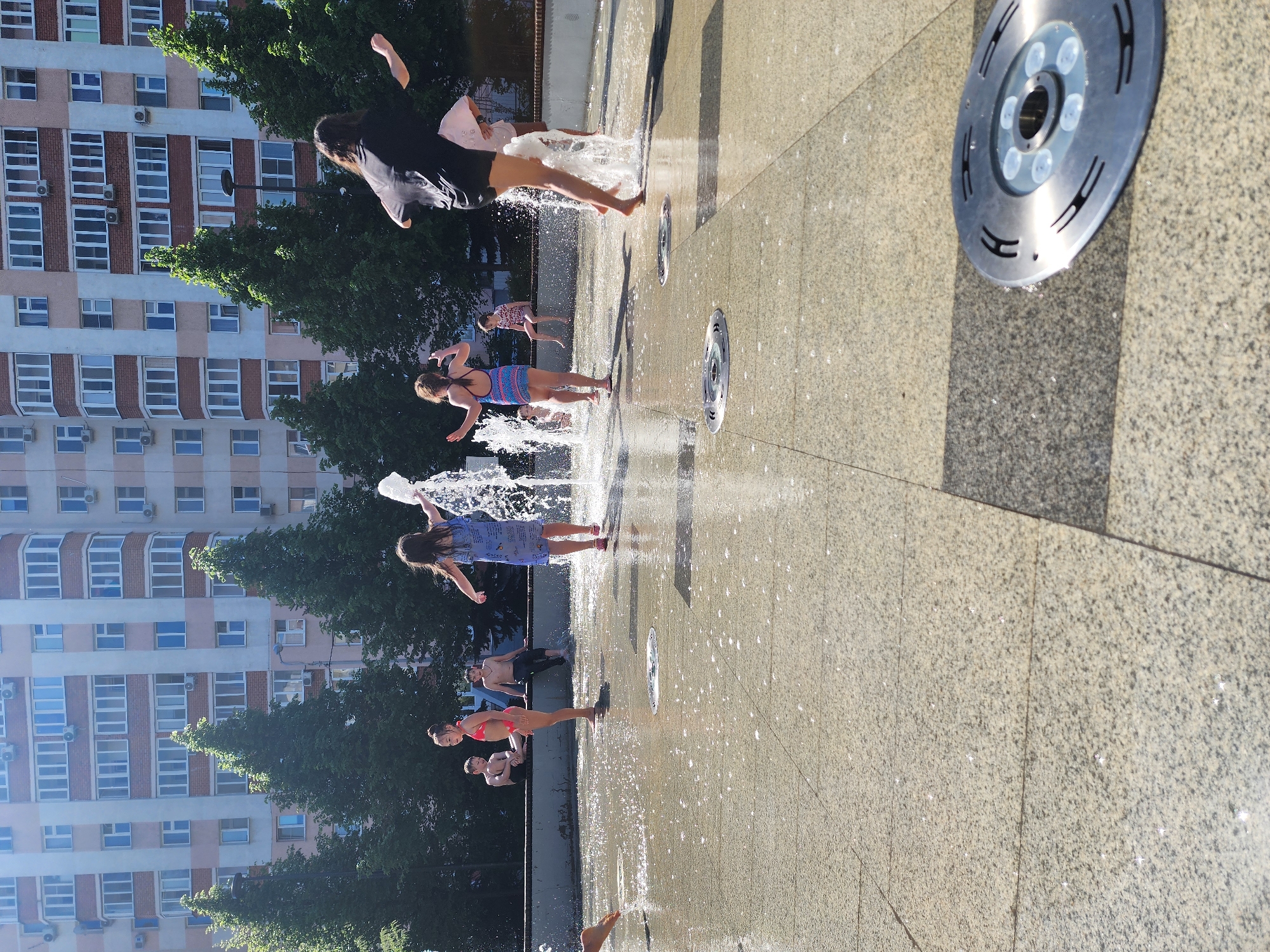 The image depicts a group of people playing in a city fountain in an urban setting. The fountain consists of multiple water jets originating from the ground, around which children are seen running and jumping. One child in a black shirt is in mid-air. Other children are wearing colorful swimwear or casual summer clothing.
The fountain's surface is a light gray stone material, with visible metal nozzles from which the water sprays. In the background, there is a multi-story residential building with numerous windows and air conditioning units attached to the facade. Green trees provide a natural contrast to the building's architectural design.
The overall scene is bright, suggesting it is daytime. The fountain appears to be a public space within a city of Unknown, Unknown.