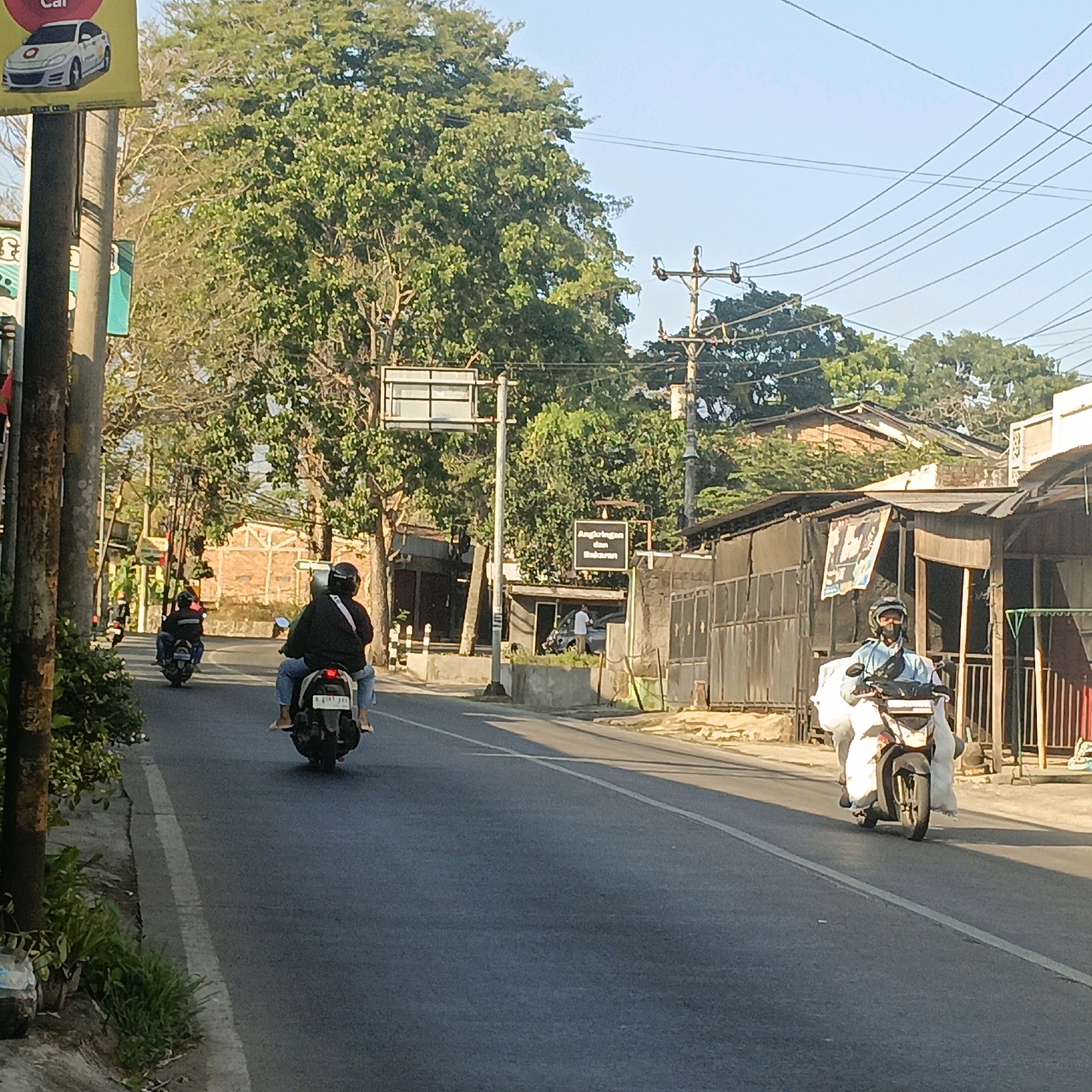 The image shows a street scene in Salatiga, Indonesia, likely during daylight hours given the bright sunlight and shadows. The road is paved and appears to be a two-lane street. There are several people riding motorcycles, moving in the same direction.
In the foreground on the right, a person wearing a light blue long-sleeved shirt, a dark helmet, and a face mask rides a motorcycle. They are carrying white bags on either side of the motorcycle. The motorcycle itself is dark-colored.
Further down the road, a person wearing a black jacket and jeans rides a motorcycle, also carrying a passenger. This motorcycle is black and has a license plate that is partially visible. Both riders appear to be wearing helmets.
On the left side of the road, a third motorcycle is visible with two riders, also wearing dark clothing and helmets.
The environment includes a sidewalk with some greenery and plants. There are poles with electrical wires overhead. Buildings are visible on both sides of the street. On the right side, there are buildings with corrugated metal roofs and a fenced area. On the left side, there is a pole with signs and a large tree. A billboard with an image of a white car and the text "Car" is also visible on the left. Another sign with Indonesian text, "Angkringan dan Bakaran," is posted on a pole near a building. The overall impression is a typical street in a city, with traffic and local businesses.