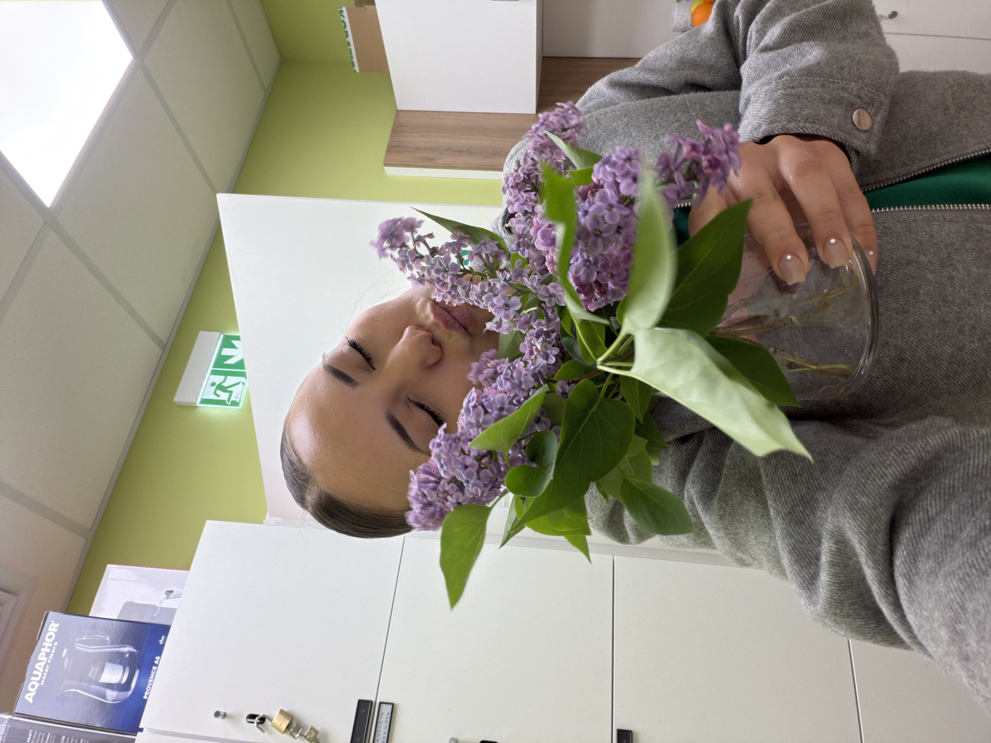 A woman is seen in an indoor environment holding a bouquet of lilac flowers in a glass vase. She is wearing a grey tweed-like jacket with a green lining visible near the zipper. The background features white cabinets, and what appears to be a green wall, an exit sign, and ceiling panels. Boxes with "AQUAPHOR Water Filters" and "PROVENCE A6" are also visible. There is not enough information to deduce the specific location in Sofia, Bulgaria.