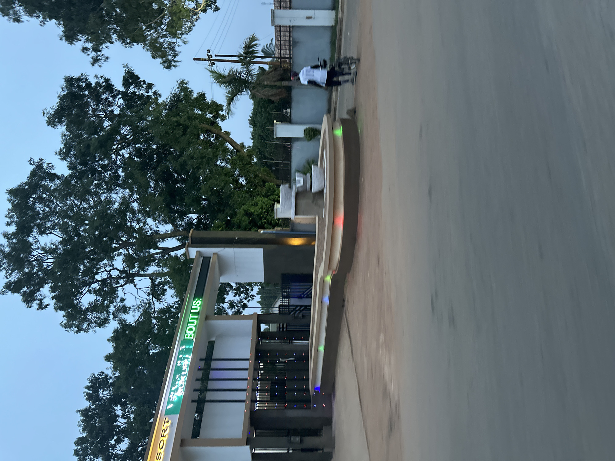 The image shows the entrance to "Bout Us Resort" located on a paved road in Bukuru, Nigeria. The entrance has a modern design with signage and decorative lighting. A person is riding a bicycle along the road in front of the resort. The area surrounding the resort features trees and vegetation. A building is visible behind the bike rider in the background. The sky is overcast.