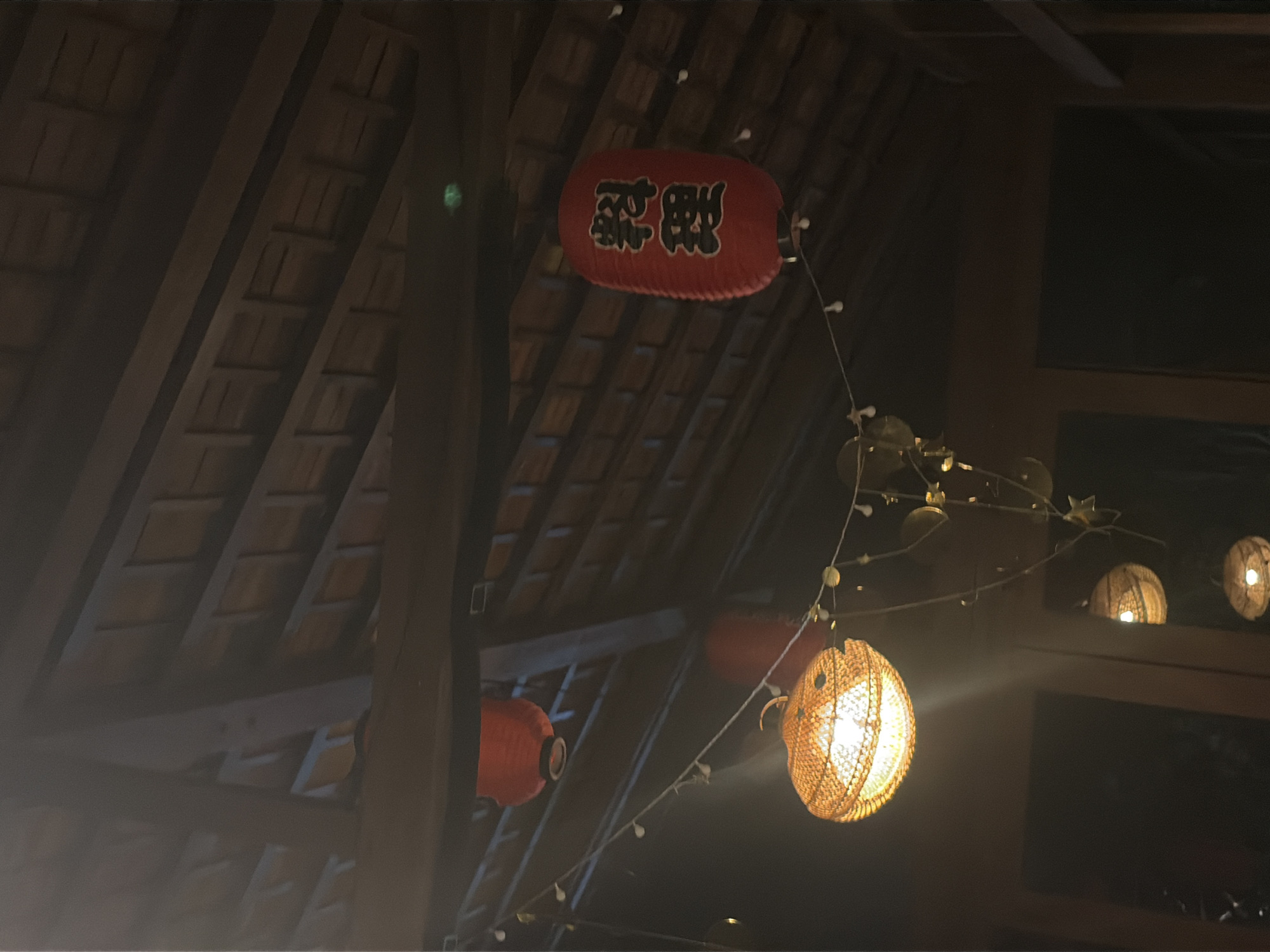 The image shows an interior view of a building with a visible wooden roof structure. Multiple lanterns are hanging from the ceiling. One prominent red lantern has Japanese characters on it, which translate to "cooking" or "cuisine." Another lantern is woven with a textured material and illuminated from within. A string of small lights runs horizontally across the scene, with decorative elements resembling leaves and stars attached. The lighting is dim, suggesting it is evening or nighttime. The building appears to be a restaurant or a similar establishment with an East Asian theme. The specific location in Kediri, Indonesia is not discernible from the image itself.