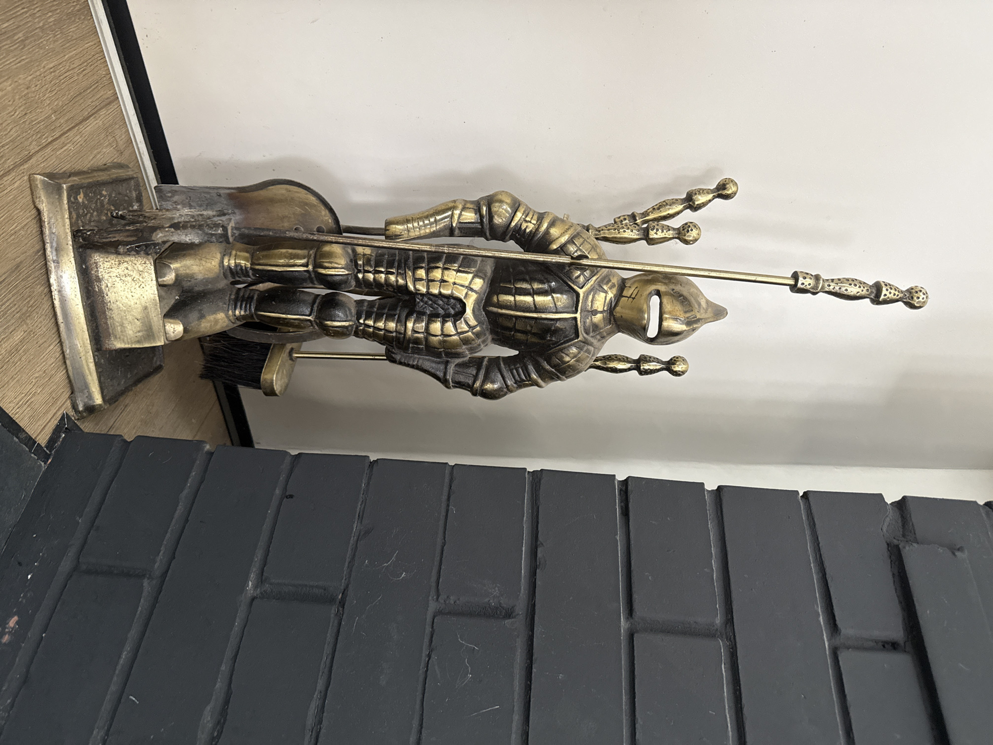 An antique brass knight-shaped fireplace tool set stands on a light wood floor next to a black brick fireplace. The knight is intricately detailed, wearing armor and holding various fireplace tools, including a poker, a brush, and a shovel. The knight's armor has a segmented design, and its face is covered by a helmet with a slit for eyes and mouth. The tools are made of brass and are integrated into the knight's design, with the poker rising from its back and the brush and shovel positioned at its sides. The fireplace is constructed of dark grey bricks and appears to be a standard size. The background consists of a plain white wall. The scene is lit by ambient light, casting soft shadows. There are no people present in the image. The location is described as Хохряки, Russia, though there is no visual information in the image to confirm this specific location.