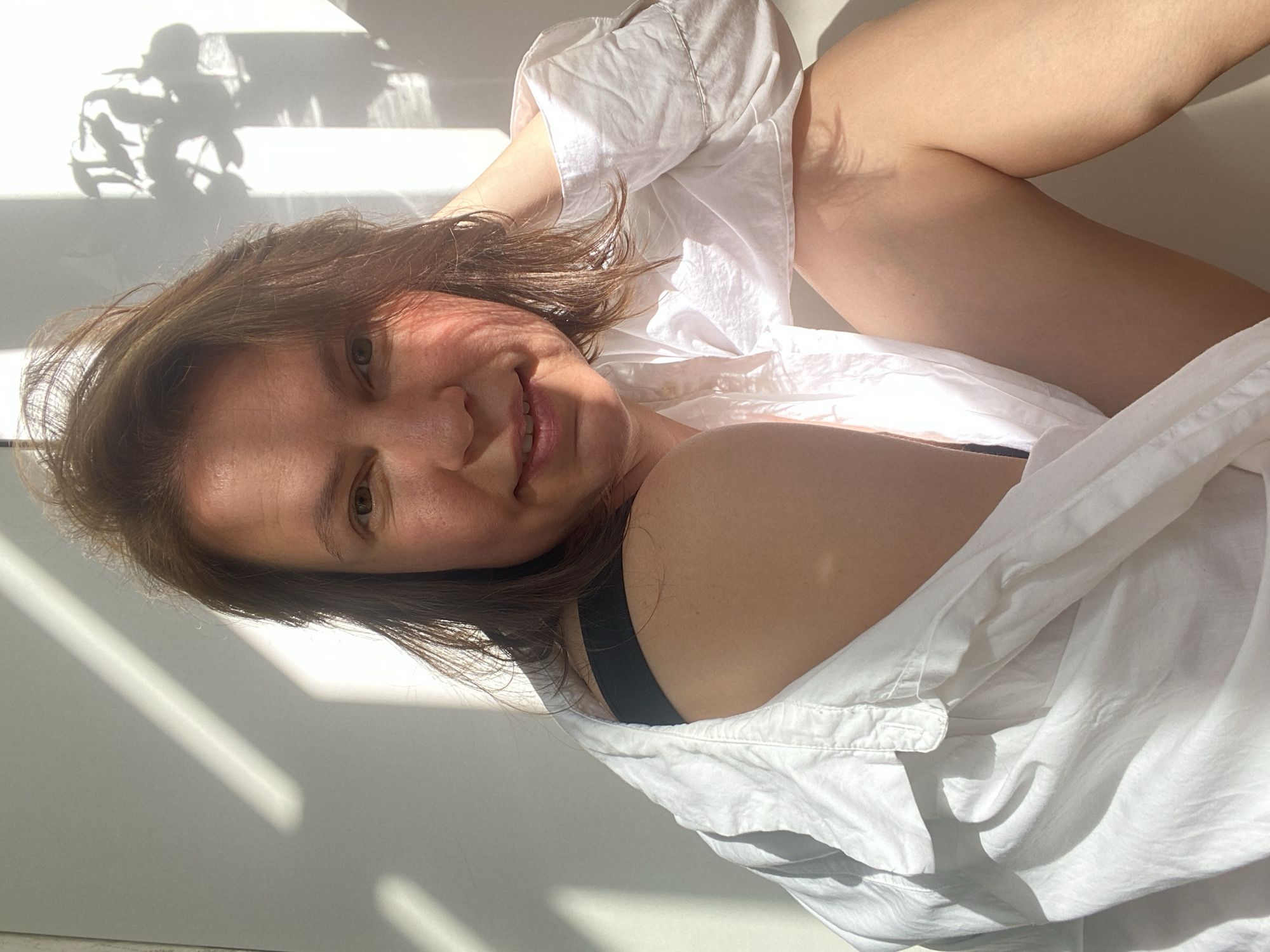 A fair-skinned woman is lying on a white surface, possibly a bed or floor, in Chaikovsky, Russia. She is wearing a black bra and a white button-down shirt. Her brown hair is disheveled, and the sunlight casts shadows across her face and the wall behind her. A plant's shadow is visible on the wall near the top left corner of the image.