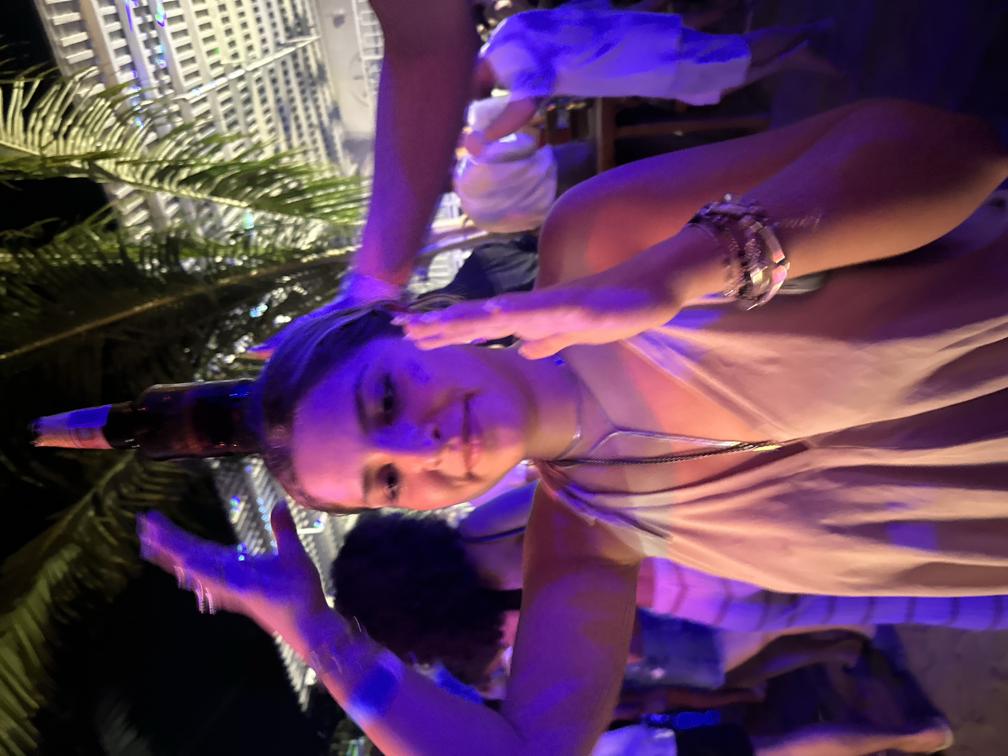 A young woman is photographed indoors, possibly at a bar or social gathering in Brisas de Zicatela, Mexico. She has a beer bottle balanced on her head. She is wearing a light-colored dress and multiple bracelets. Other people are visible in the background, including someone wearing white clothing. Palm trees and a building with lights are visible outside. The image appears to have a blue and purple hue due to the lighting.