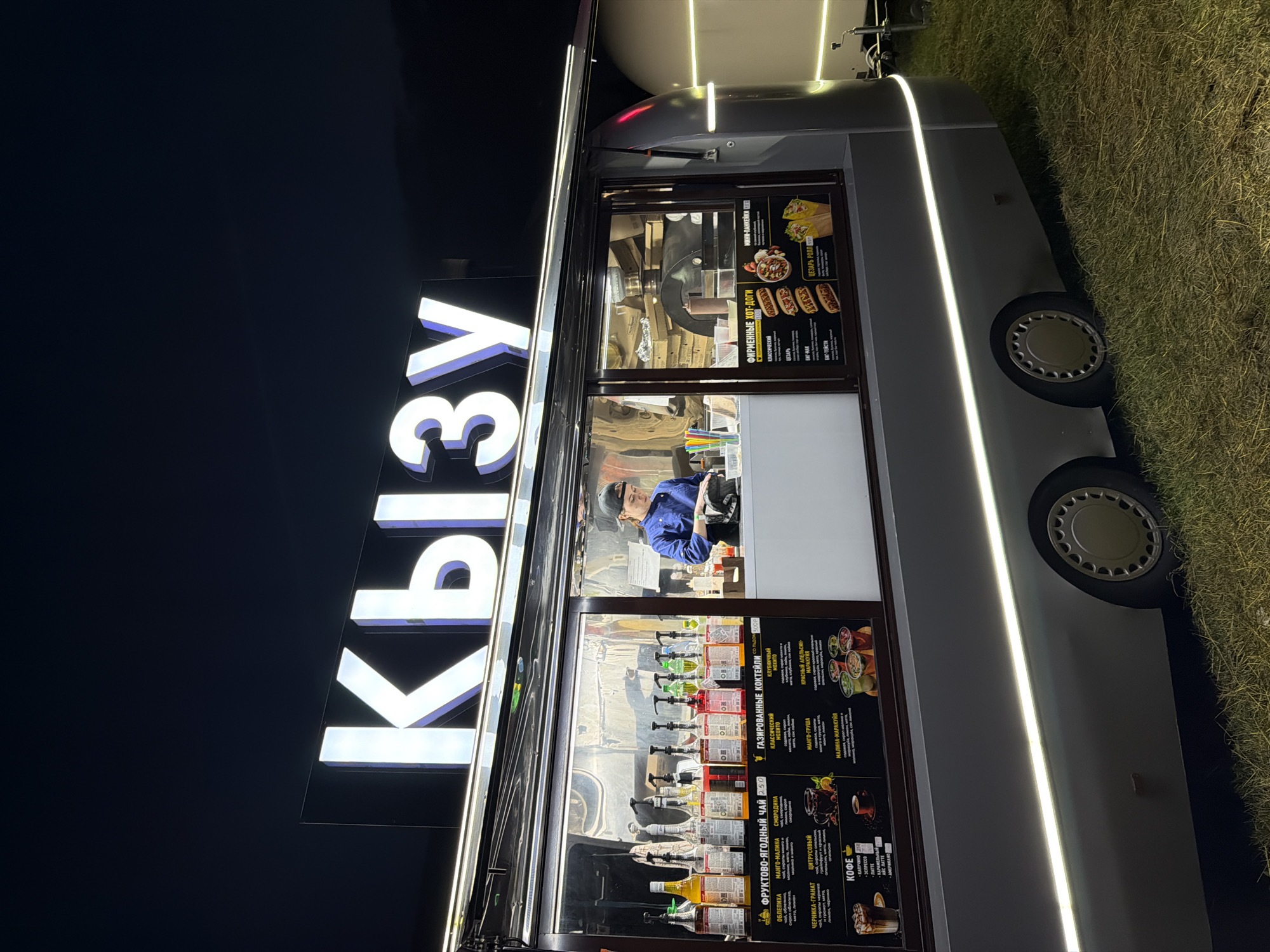 This is a vertical, eye-level, outdoor shot of a food truck at night. The food truck is illuminated by bright white LED strip lights outlining its body and a large, illuminated sign that reads "КЫЗУ" in white, block letters. The truck is parked on a grassy area.
Inside the truck, a person wearing a blue shirt and a baseball cap is visible behind a counter, appearing to be an employee. The windows of the food truck display menus with various food and drink options. Visible text includes titles such as "ФРУКТОВО-ЯГОДНЫЙ ЧАЙ" (Fruit-Berry Tea), "ГАЗИРОВАННЫЕ КОКТЕЙЛИ" (Sparkling Cocktails), "ФИРМЕННЫЕ ХОТ-ДОГИ" (Signature Hot Dogs), and "КОФЕ" (Coffee). Specific items mentioned on the menus include "Облепиха" (Sea Buckthorn), "Манго-Малина" (Mango-Raspberry), "Черника-Гранат" (Blueberry-Pomegranate), and "Цезарь Ролл" (Caesar Roll).
The overall scene suggests a lively, nocturnal food service operation. The lighting creates a vibrant atmosphere, and the presence of the menu items indicates a focus on beverages and fast food. The grass suggests an outdoor event or a stationary roadside location. The time of day is clearly night due to the dark background and artificial lighting.