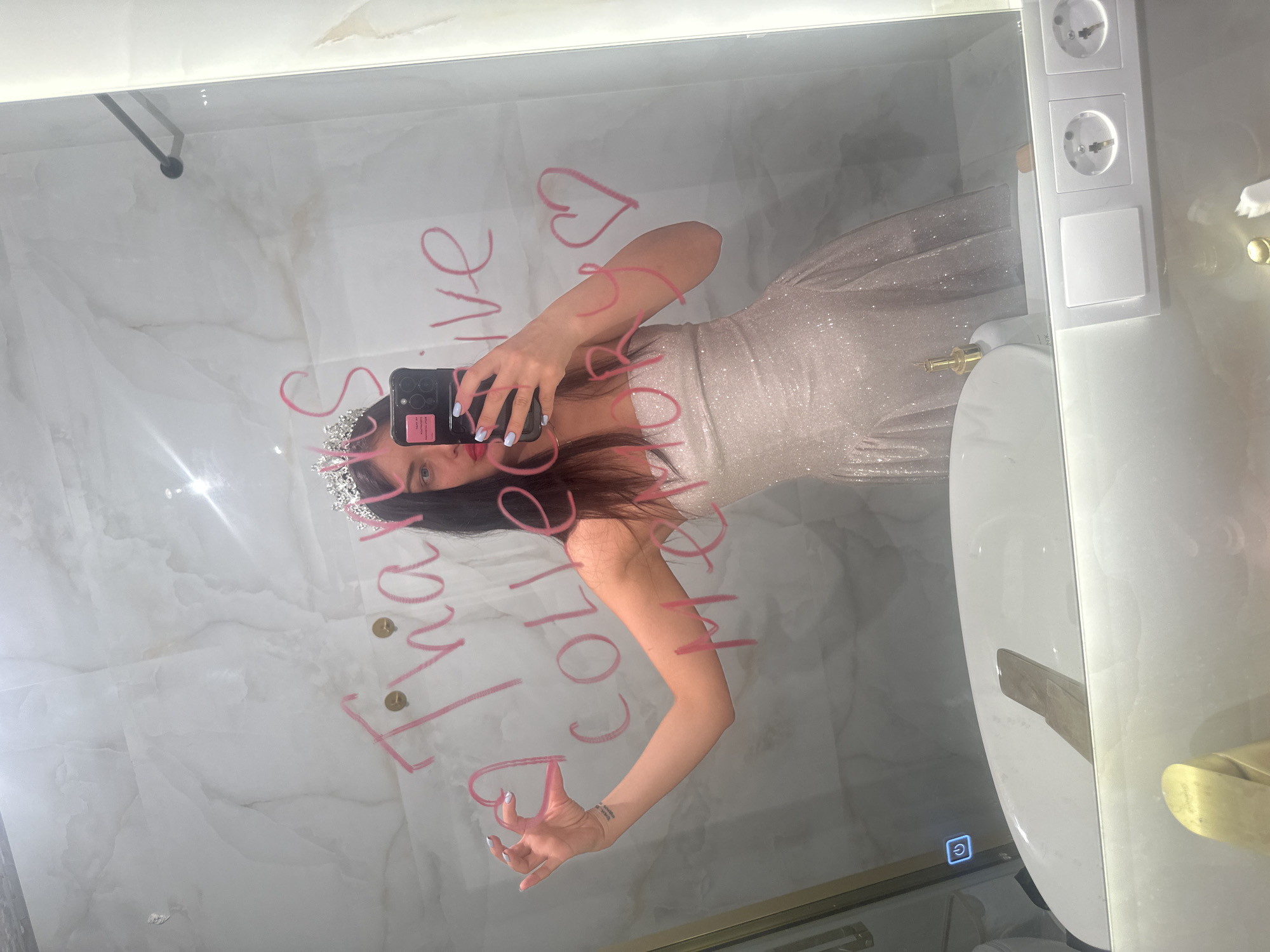 The rotated image captures a mirror selfie taken by a woman in what appears to be a modern bathroom or dressing room in Moscow, Russia.
**Subjects:** The primary subject is a woman, reflecting in the mirror, holding a black smartphone with a textured case. She has long, dark hair, light-colored eyes, and her nails are painted a light blue or lavender shade. She is wearing a sparkly, light-colored, strapless formal dress, possibly white or pale pink, and a tiara on her head. A small tattoo is visible on her left forearm.
**Objects:** The central object is the large mirror, serving as the surface for the selfie. Written in pink, likely lipstick or a marker, are the words "Thank you, Colleen" followed by a heart symbol. To the right of the mirror, a white vertical panel features two European-style electrical outlets (Schuko sockets) and two square switches. Below these, a gold-toned faucet or fixture is partially visible. At the bottom left edge of the mirror's frame, a blue illuminated power symbol is present, suggesting a lighted mirror feature. The wall reflecting behind the woman appears to be covered in a light-colored, possibly white marble or veined tile pattern. The ceiling above the mirror shows some minor discoloration or possible water damage.
**Scene and Setting:** The setting is an indoor space, likely a contemporary and well-lit bathroom or dressing area, indicated by the mirror, marble-like surfaces, and electrical fixtures typical of such spaces.
**Activity:** The woman is actively taking a mirror selfie, holding her phone to capture her reflection.
**Notable Details:** The image is rotated 90 degrees clockwise. The formal attire (sparkly dress and tiara) suggests a special occasion or celebration. The European electrical outlets are a subtle location cue, consistent with the specified location context of Moscow, Russia. The "Thank you, Colleen" message on the mirror adds a personal and appreciative touch. The time of day is not discernible due to artificial lighting, and weather is irrelevant for this indoor scene. Emotions conveyed appear celebratory or thankful, fitting the context of the dress and message.
**Visible Text:** "Thank you, Colleen" and a heart symbol are written in pink on the mirror. A blue illuminated power symbol is on the mirror frame.