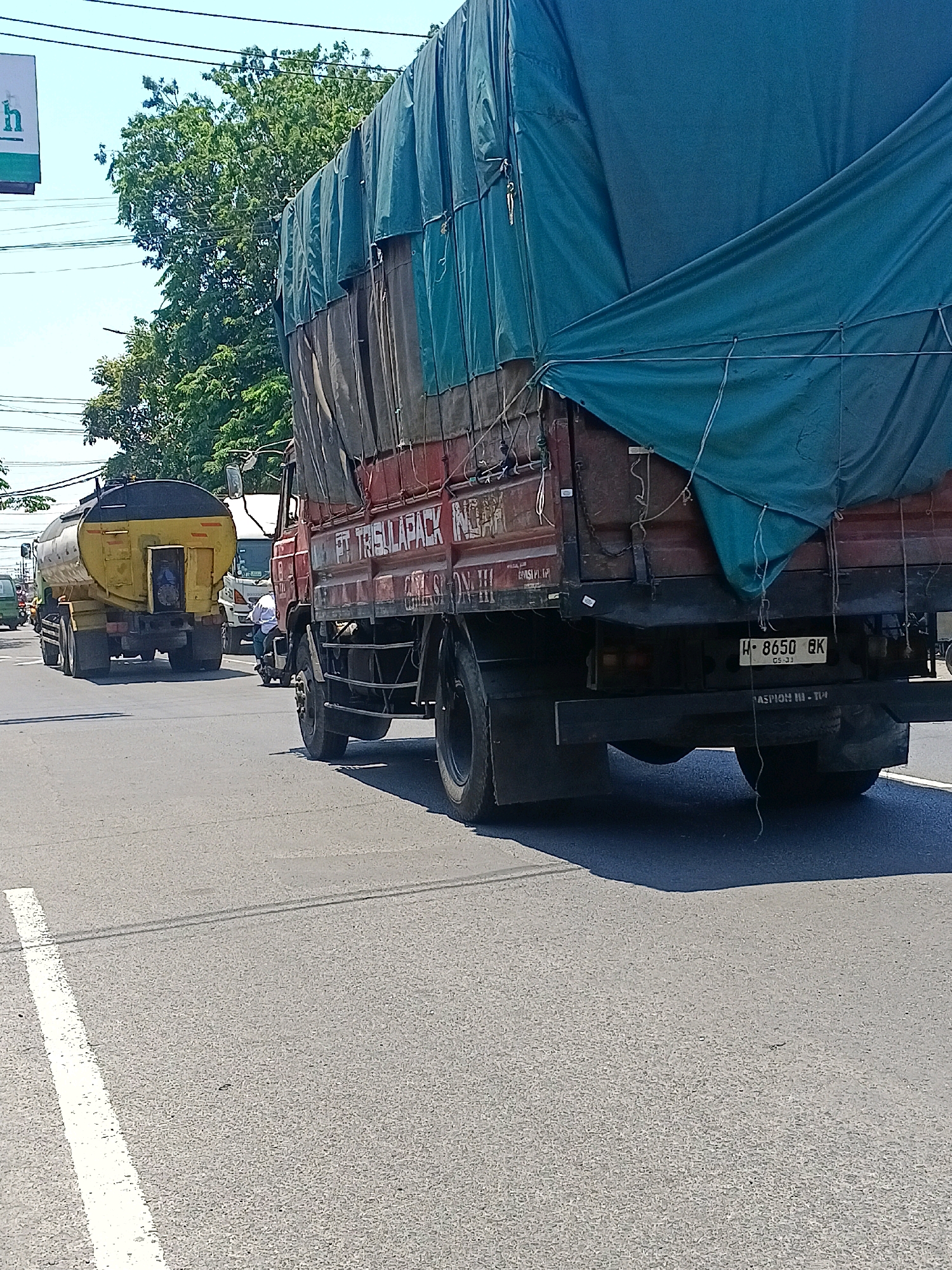 In Kalijaten, Indonesia, on a sunny day, a red truck with a green tarp covering its cargo is seen on the right side of the road. The truck's license plate reads "H-8650-BK". On the left side of the road, a yellow tanker truck is driving, also on the asphalt. In the background, there are trees and power lines. The scene depicts traffic on a road with the typical urban elements of the Indonesian city.