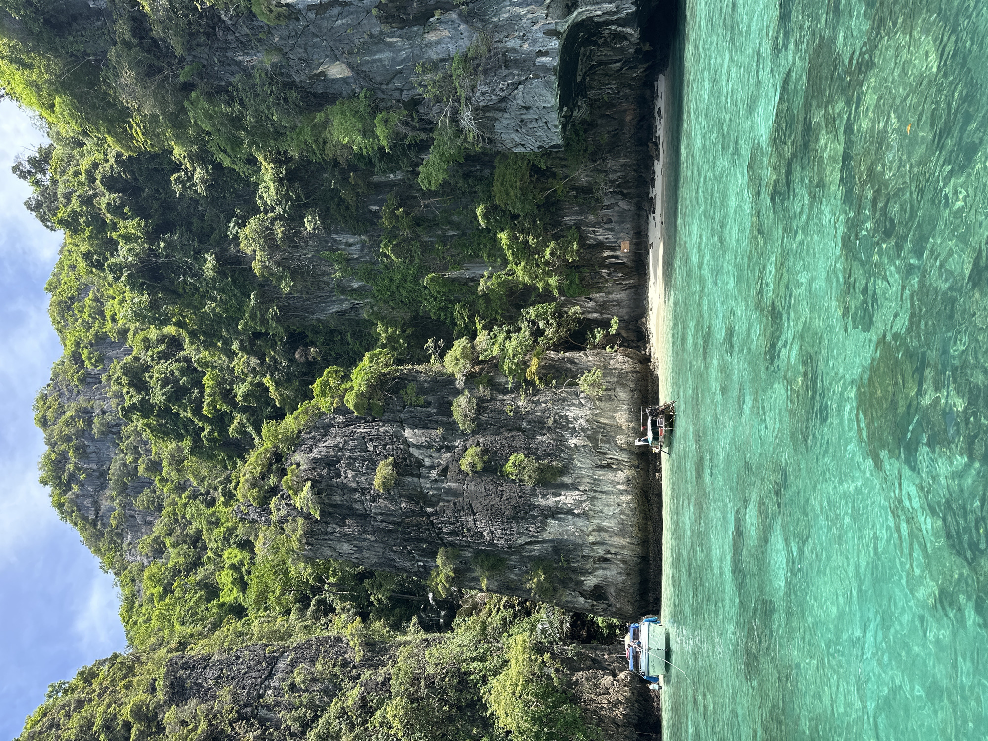 The image depicts a scenic view of limestone cliffs covered in lush green vegetation, characteristic of the islands surrounding Ao Nang, Thailand. Crystal-clear turquoise water fills the foreground, with two boats anchored offshore. One boat, a white speedboat, is closer to the left, with its anchor rope visible. To the right, a smaller, darker wooden boat, likely a longtail boat, is moored. The background consists of towering cliffs with sheer rock faces, interspersed with dense foliage and trees clinging to the rocky surfaces. A small, sandy beach is visible nestled between the cliffs, with a signpost standing on it. The sky above is partly cloudy with patches of blue. The overall impression is one of natural beauty and tranquility.