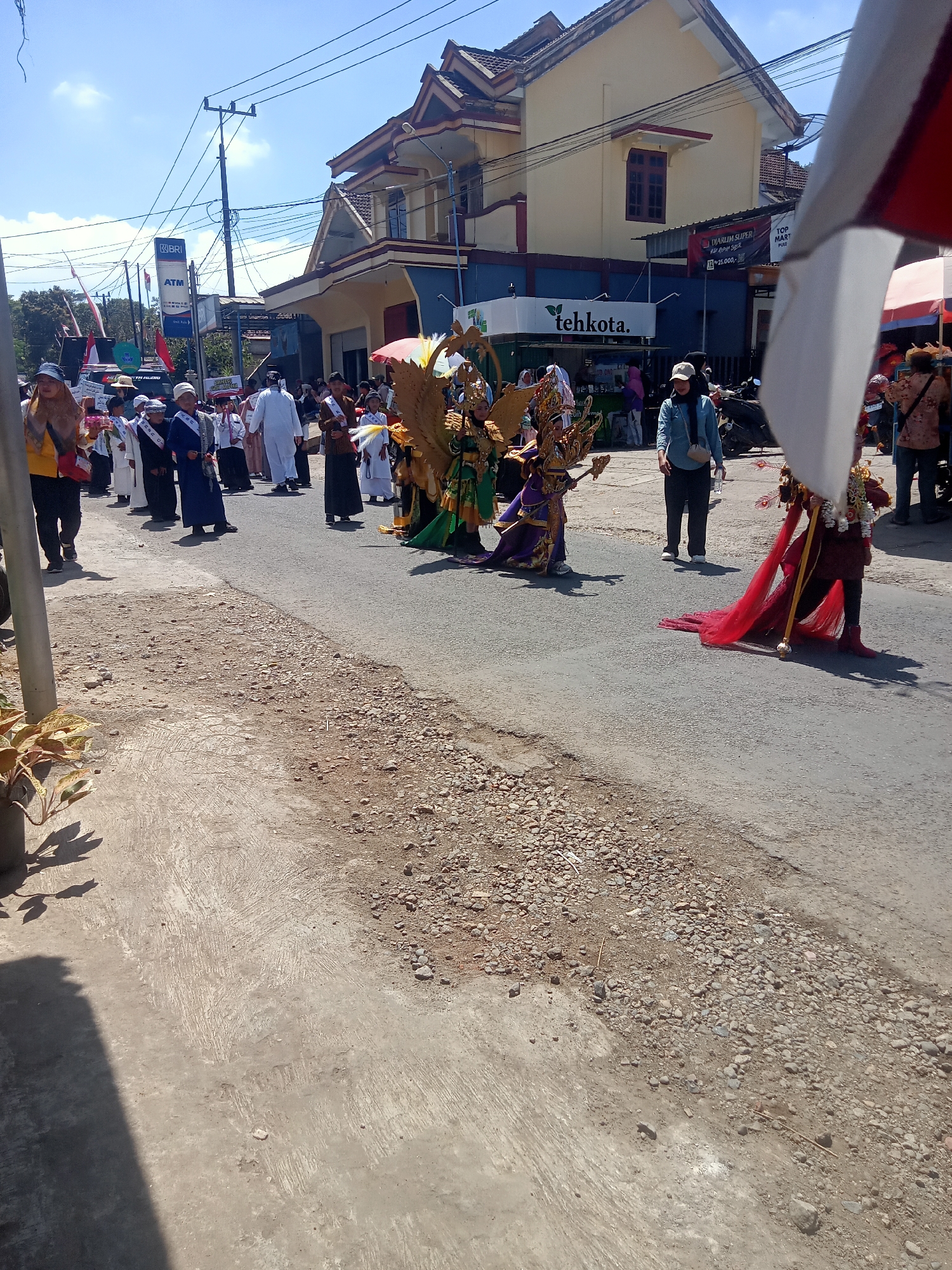 In the city of Pule, Indonesia, a street parade is underway under a bright, sunny sky. A procession of people, including many children dressed in white, walk along an asphalt road. Some children wear sashes with text. In the background, a yellow and blue building with a red-tiled roof stands. Signs on the building include "tehkota." and "DIARUM SUPER". To the left of the procession, a sign for "BRI" and "ATM" is visible on a utility pole. Further down the street, individuals in elaborate costumes, one adorned with large golden wings and another in a red cape, participate in the parade. A person wearing a blue shirt and a beige cap stands to the side, observing. The street is bordered by concrete on the left and a mixture of gravel and dirt on the right.