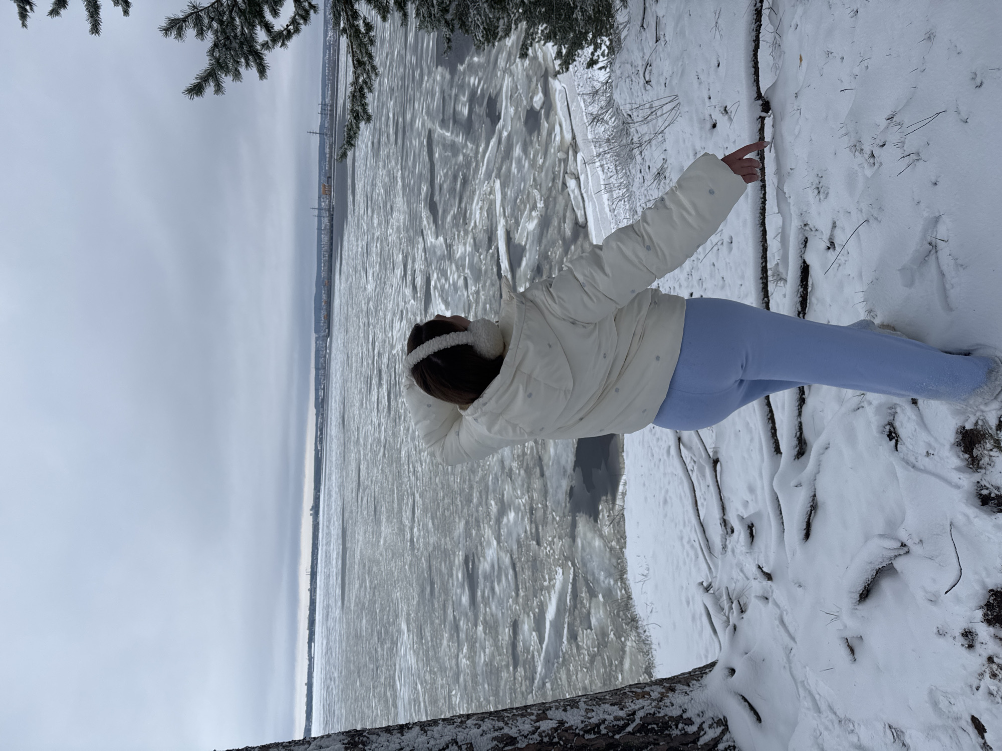A woman in a white jacket and light blue pants stands on a snowy bank, pointing toward a wide, icy river. The river is covered in broken ice floes. In the distance, a cityscape with tall structures is visible through a hazy sky. The location is likely Noviy, Russia. Branches of snow-covered trees frame the top and bottom of the scene. The woman is wearing white earmuffs.