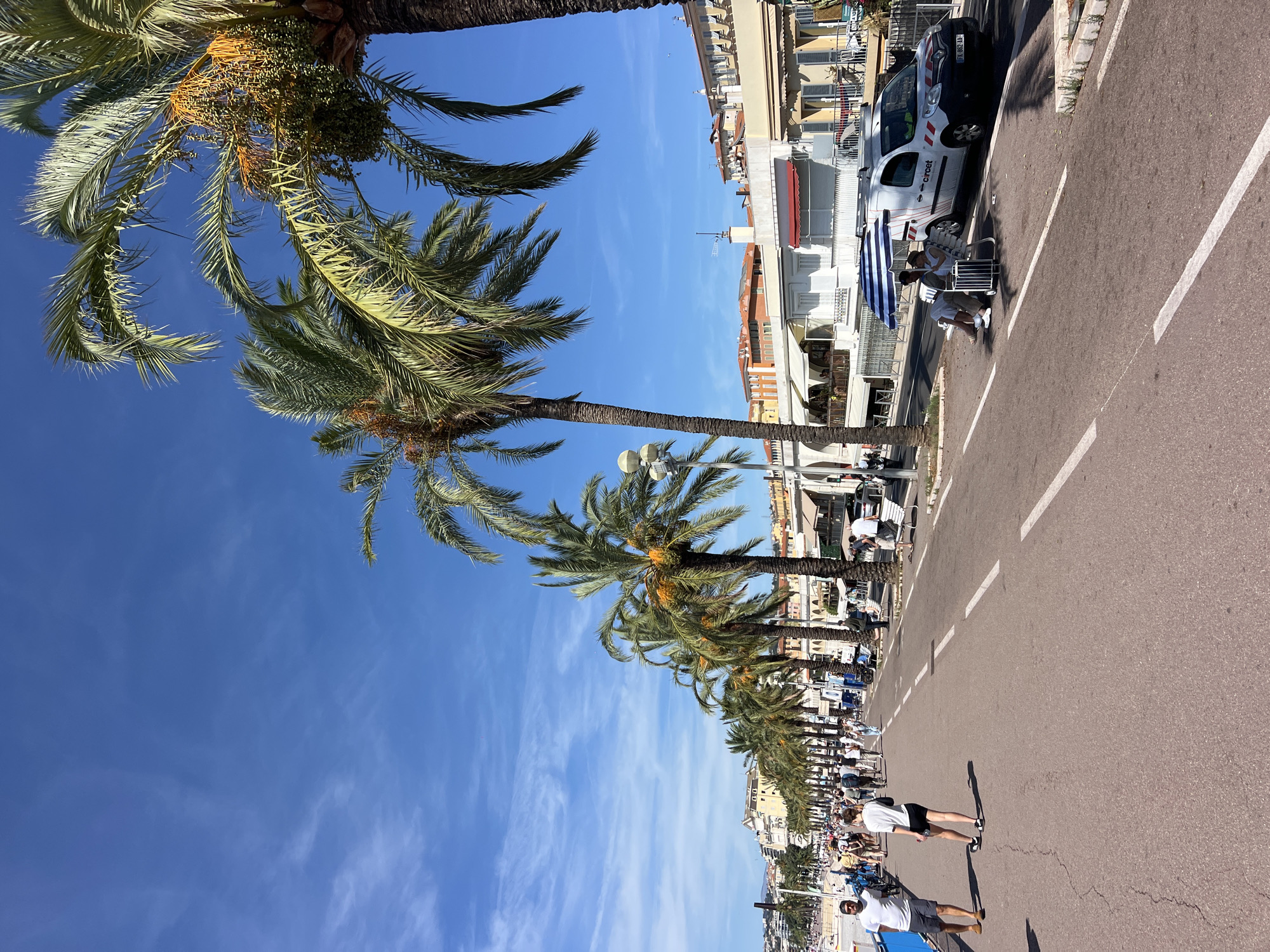 The image shows a sunny day on the Promenade des Anglais in Nice, France. A tree-lined walkway borders a busy road where cars and people are present. Palm trees are abundant, with some bearing fruit. The buildings in the background are typical of the French Riviera, with awnings and balconies. Two people are sitting under an umbrella on a bench near a white van with "circet" written on its side. The sky is a clear blue with some wispy clouds.