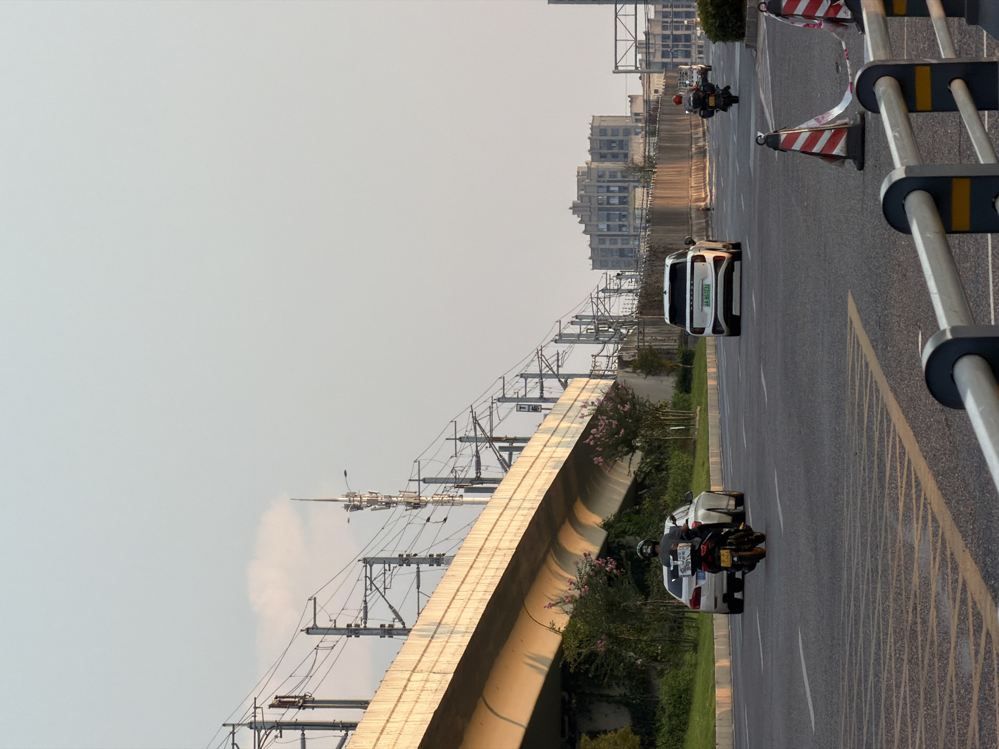 This is a vertical, outdoor, medium shot of a road in Jiangjin, China, during what appears to be late afternoon or early evening. The sky is a pale, clear blue with a faint, wispy cloud. In the foreground, the asphalt road features yellow lane markings. A silver sedan is visible in the left lane, and a black motorcycle with a rider in a helmet and dark clothing is in the right lane, following the car.
Further down the road, a white SUV is in the right lane, and a motorcycle with a rider wearing a red helmet and a dark backpack is in the right lane. Traffic cones with red and white stripes, some with torn tape, are placed on the right side of the road. A silver metal guardrail runs along the bottom of the image.
In the background, an elevated concrete structure, possibly an overpass or elevated train line, dominates the left side. It is supported by pillars and adorned with numerous electrical wires and poles. Green foliage and some pink flowering bushes are visible beneath the overpass.
To the right of the overpass, a tall, multi-tiered antenna or communication tower stands against the sky. Beyond the road, a cluster of buildings with a distinctive, stepped architectural style can be seen in the mid-ground. Further in the distance, more urban structures are visible. The overall impression is one of an urban environment with infrastructure for transportation and communication.