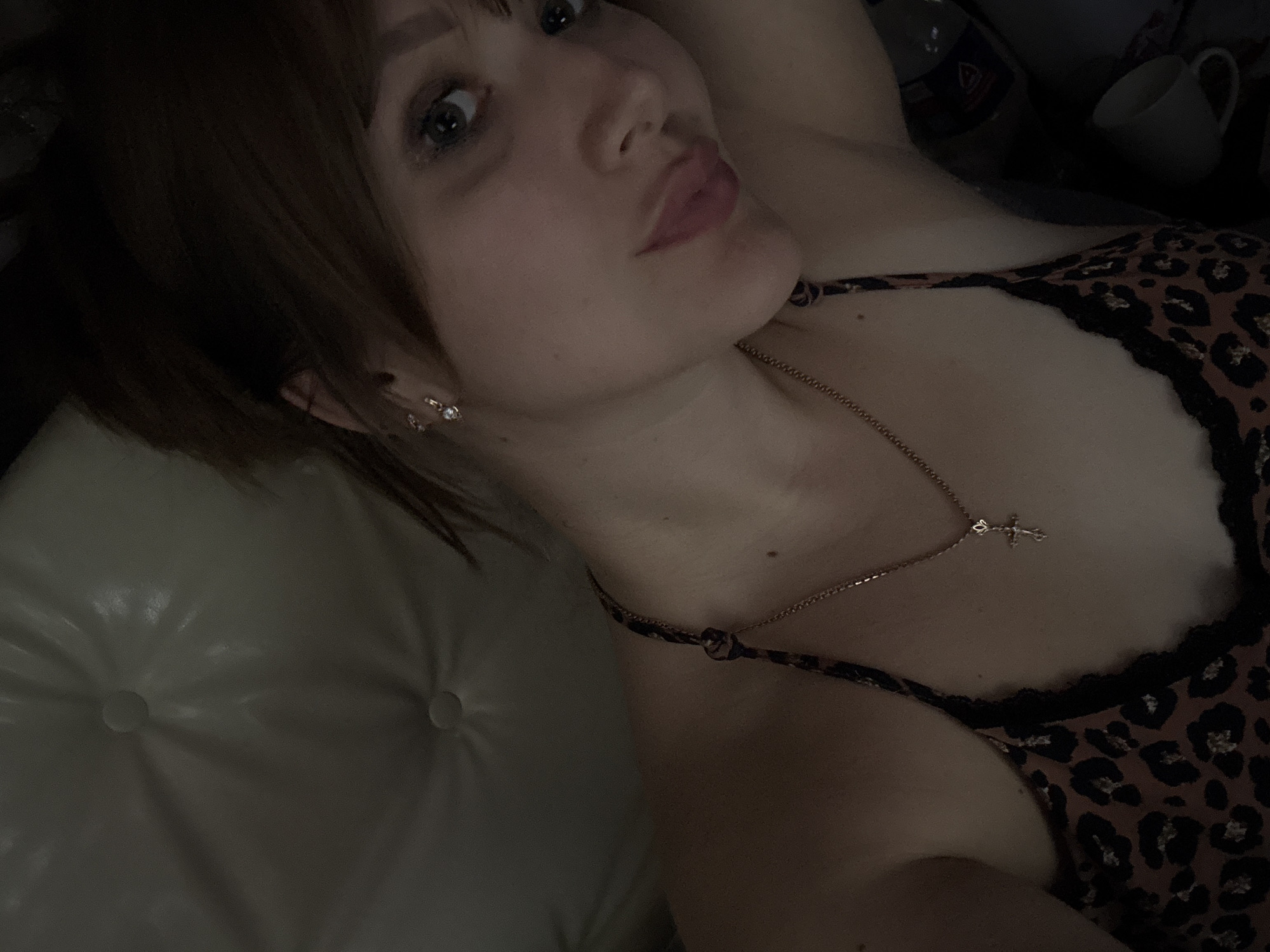 The image is a close-up, selfie-style photograph of a young woman lying down. She has brown hair, styled with bangs, and is wearing a leopard-print camisole with black lace trim and a thin gold chain necklace with a cross pendant. She is also wearing small stud earrings. The background appears to be a light-colored, tufted surface, possibly a sofa or bed. The lighting is dim, creating shadows and highlighting the woman's face and upper body. The image does not contain any discernible elements that would indicate a specific location within Chaikovsky, Russia, or any other city.