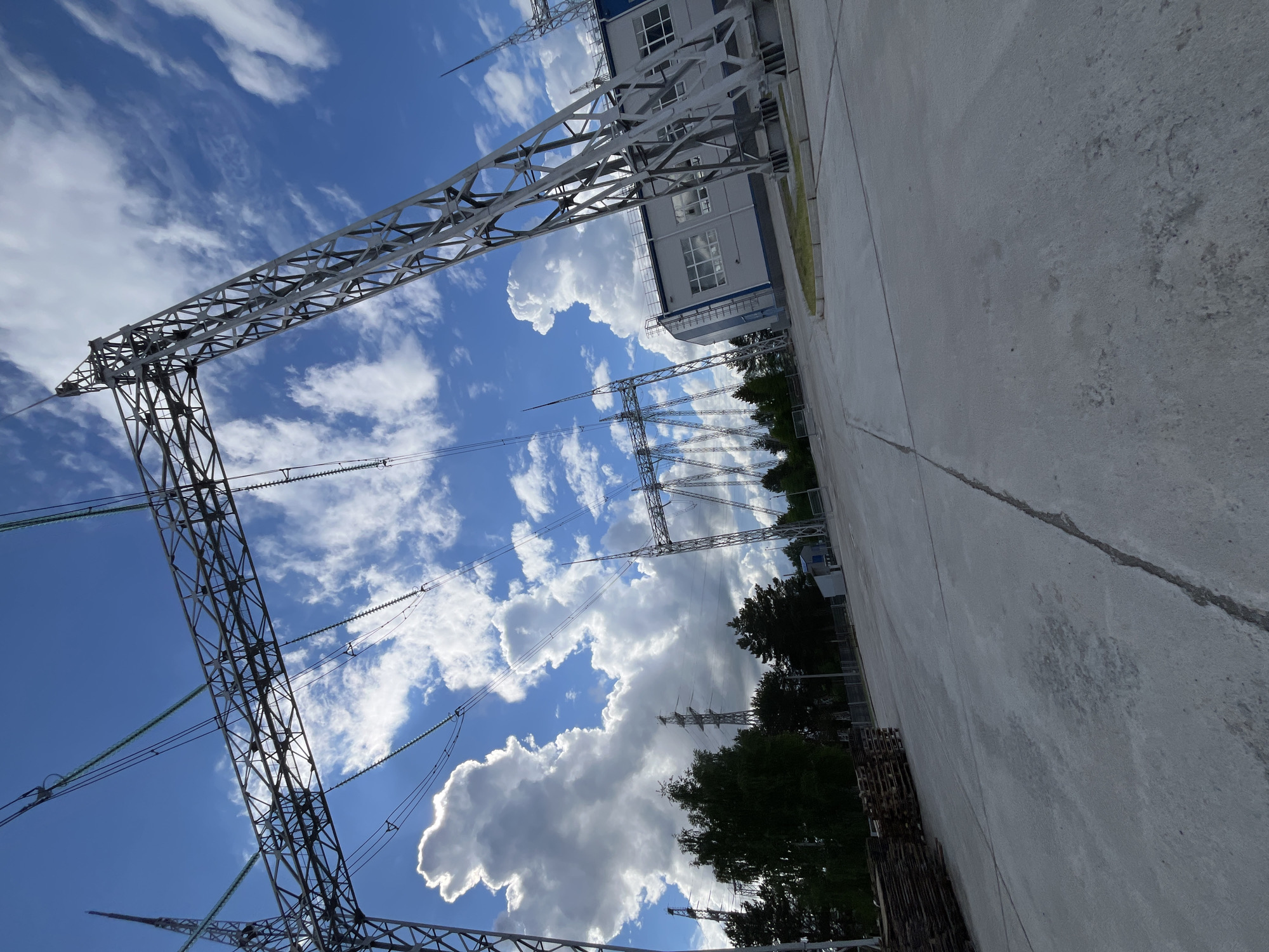 The image, oriented on its side (rotated 90 degrees clockwise), captures a bright daytime scene at what appears to be an electrical substation or power generation facility in Chaikovsky, Russia. The dominant feature is the expansive sky, a vibrant blue with scattered white, fluffy cumulus clouds. Many clouds have intensely bright, sunlit edges, indicating direct sunlight and clear weather.
In the left and central parts of the frame, several large metallic lattice transmission towers (pylons) are prominent, reaching towards the sky. These towers support numerous power lines and insulators, which are visible against the blue sky, some appearing to have a greenish tint.
Towards the upper right, a modern, light-colored building with multiple windows and a blue trim is visible, likely an administrative or control building for the facility. Below the building and extending into the mid-ground, dark green trees provide a natural element against the industrial backdrop, partially obscuring the distant landscape.
The foreground and right side of the image are dominated by a wide expanse of concrete or asphalt ground, showing some cracks and stains, typical of an industrial area. In the lower right, stacks of wooden pallets are visible on the ground. No people or activities are visible, and there is no visible text. The overall impression is one of a functional, industrial setting under a bright, clear sky.