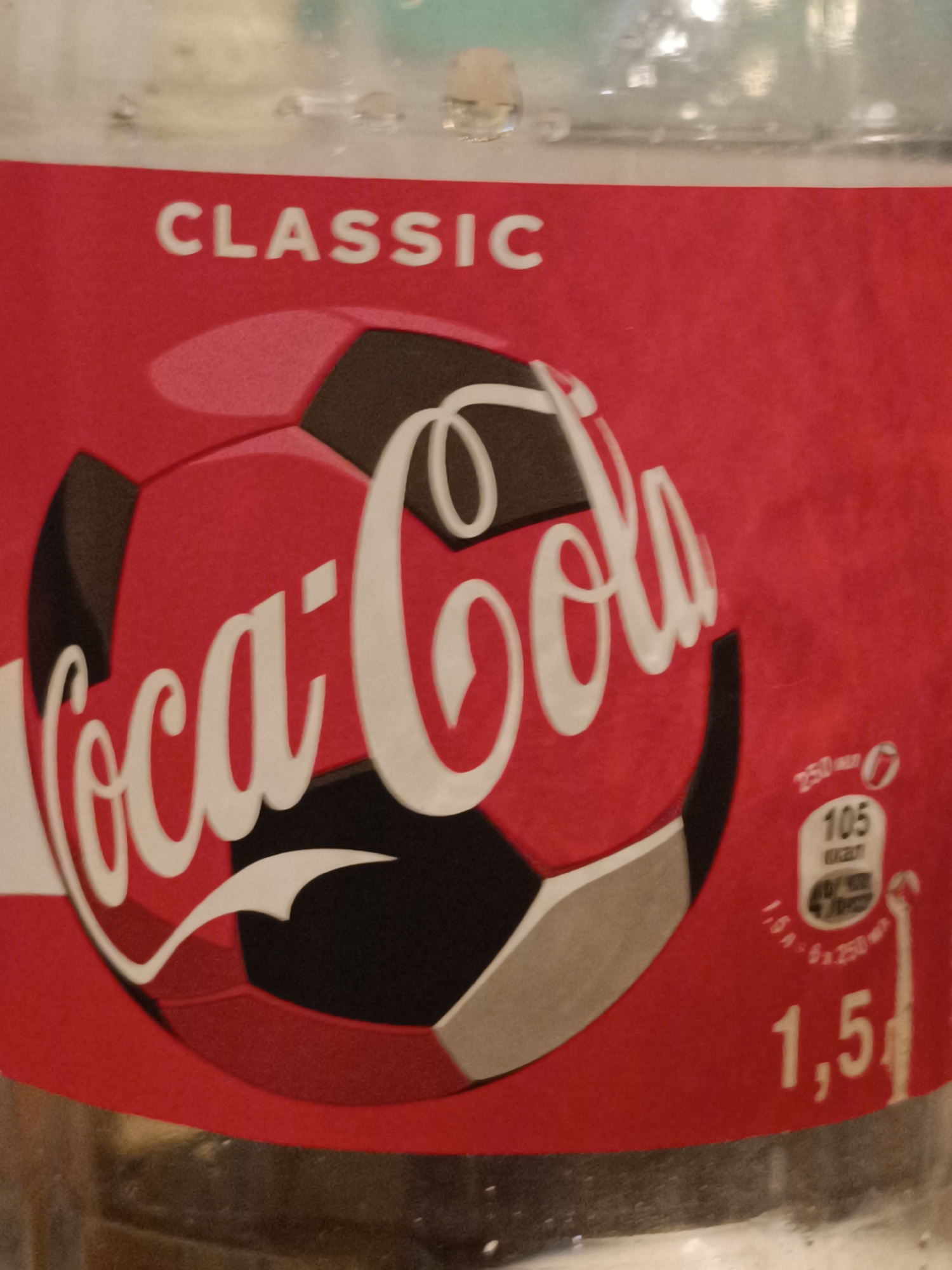 The image shows a close-up of a Coca-Cola bottle. The bottle has a red label featuring the Coca-Cola logo in white script, superimposed on a graphic of a soccer ball. Above the logo, the word "CLASSIC" is printed in white capital letters. To the right of the logo, there is nutritional information indicating "250 ml - 6" and "105 kcal". Below this, "1.5L" is printed in large white numbers, along with smaller text and a graphic showing a bottle outline with measurements. The background is blurry and appears to be the interior of a refrigerated case or display, with hints of other bottles and reflections. There are no people visible in the image, nor is there any indication of the city of Taraz city administration, Kazakhstan.