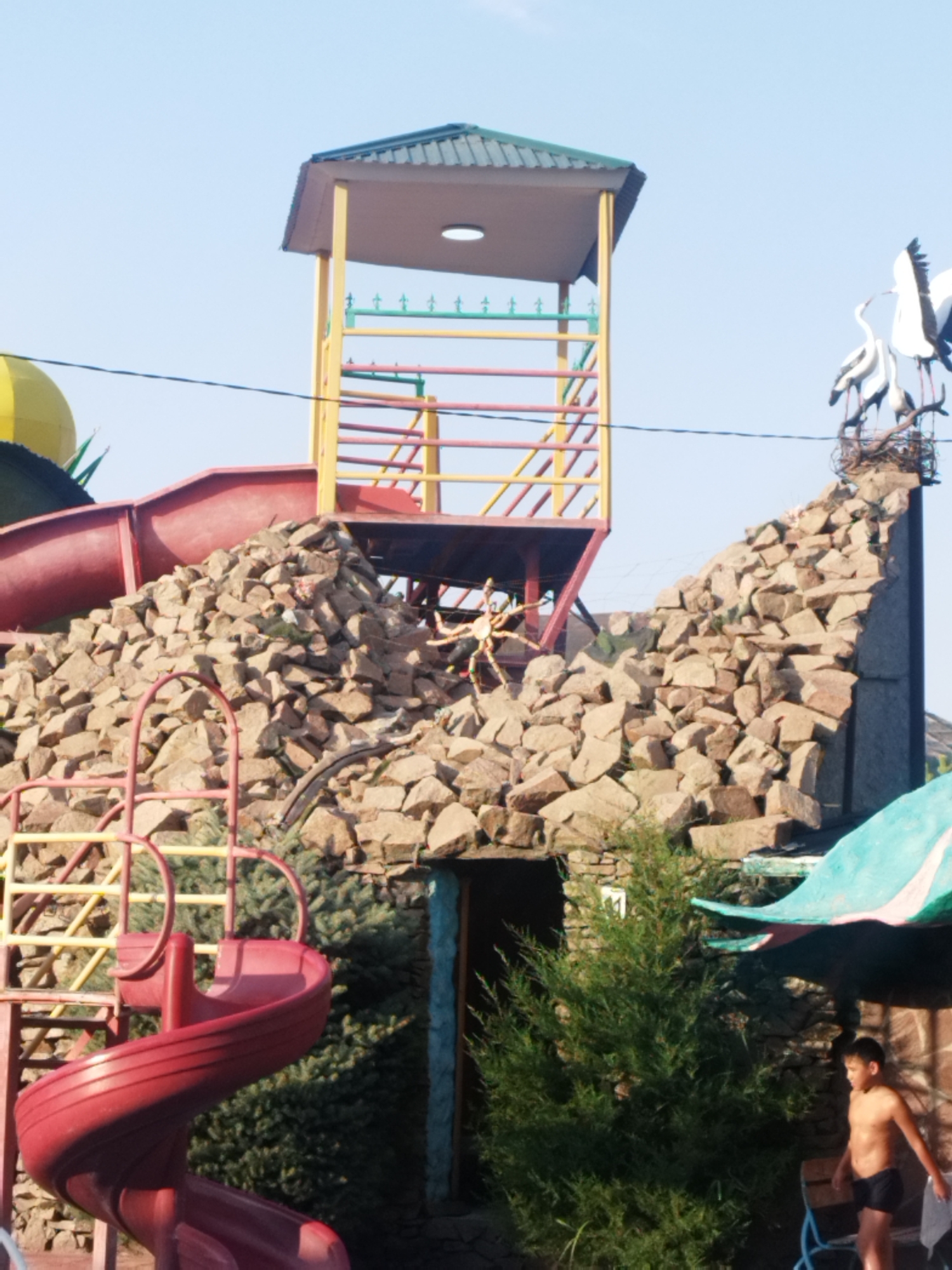 The image depicts a vibrant water park attraction with a towering slide structure in the city of Shymkent, Kazakhstan. The main structure features a multi-level tower with colorful railings (yellow, green, and red) supporting a roofed observation deck. A prominent red spiral slide curves downwards from a higher level, disappearing behind a large pile of rough, tan-colored stones. The stones are arranged to resemble a natural rock formation, with some greenery peeking through.
To the right of the main structure, a large, decorative nest with two white stork figures is perched atop a rocky facade. Below this, a young boy, appearing to be around 8-10 years old, stands shirtless in dark swim trunks. He is holding a white object in his right hand and is positioned near a wooden bench and a blue chair. A large, green, dragon-shaped canopy extends from the right side of the image, partially obscuring a stone-faced building entrance. The environment suggests a sunny day with a clear blue sky overhead. The overall scene is one of outdoor recreation and entertainment.