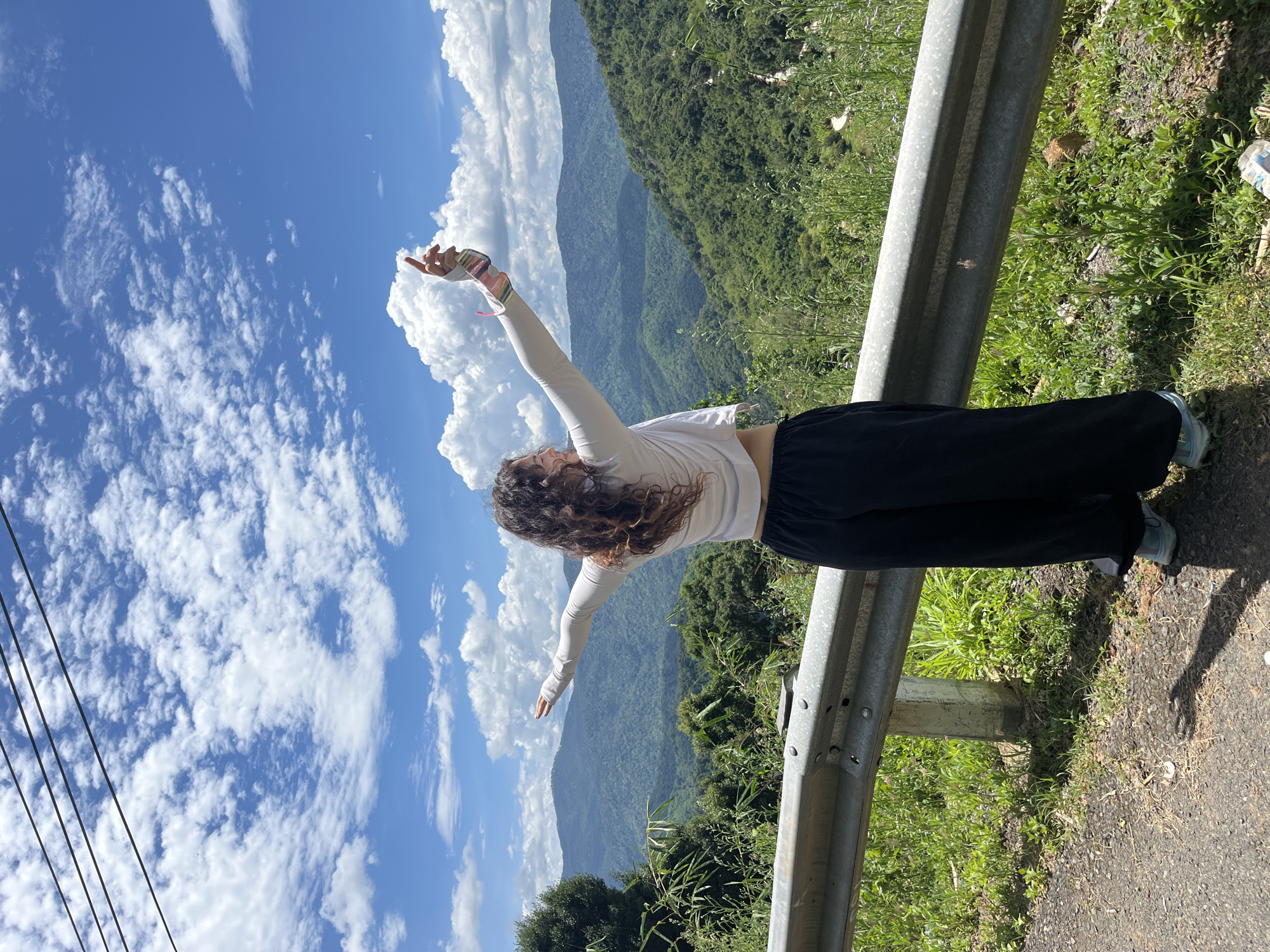 A young woman with curly brown hair is standing on the side of a road, facing away from the camera, with her arms outstretched and raised towards a bright blue sky filled with white, fluffy clouds. She is wearing a white long-sleeved top, black loose-fitting pants, and light blue athletic shoes. In her right hand, she holds a colorful object, possibly a phone or a small device. The road is paved with asphalt and bordered by a metal guardrail. Lush green vegetation, including trees and grasses, grows on the embankment behind the guardrail. In the background, rolling hills covered in dense green forest stretch as far as the eye can see under the expansive sky. The scene suggests a location with natural beauty and an open, elevated viewpoint, consistent with mountainous terrain. This scene is located in the city of Son Dien, Vietnam.
