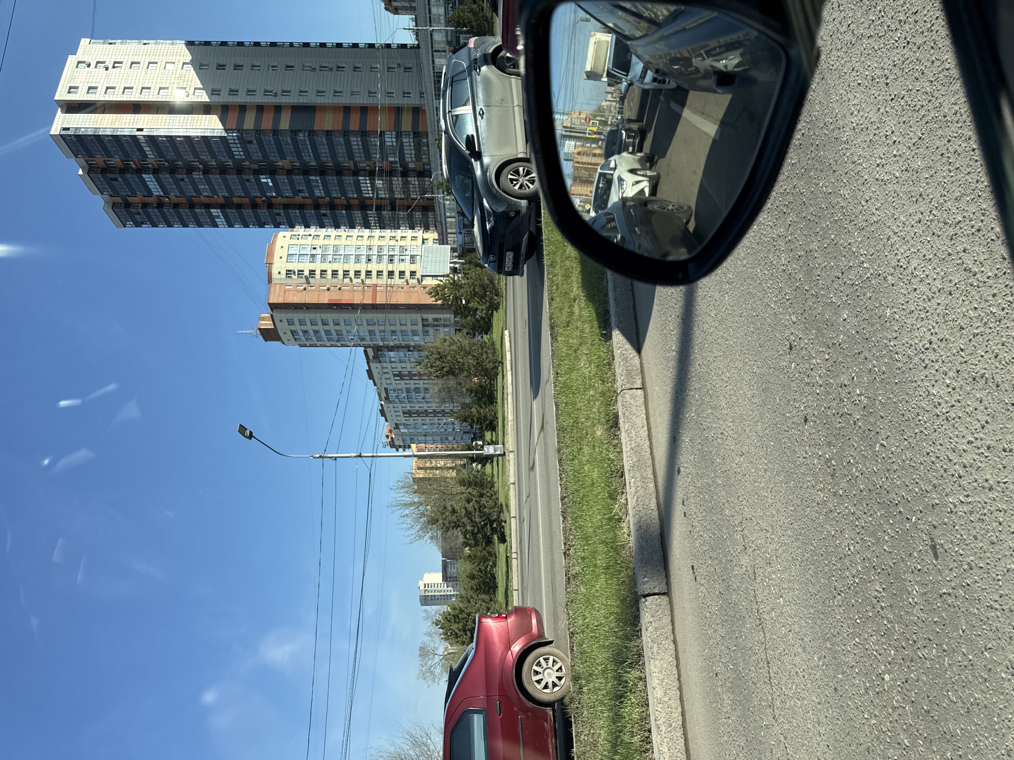 The image shows a street scene in Krasnoyarsk, Russia, captured from inside a vehicle. A gray car is parked alongside the road. The buildings, trees, and utility poles are visible in the reflection in the car's side mirror. There are tall apartment buildings in the background under a clear blue sky. A red vehicle is parked closer to the foreground. The road is asphalt with grass strips on the side.