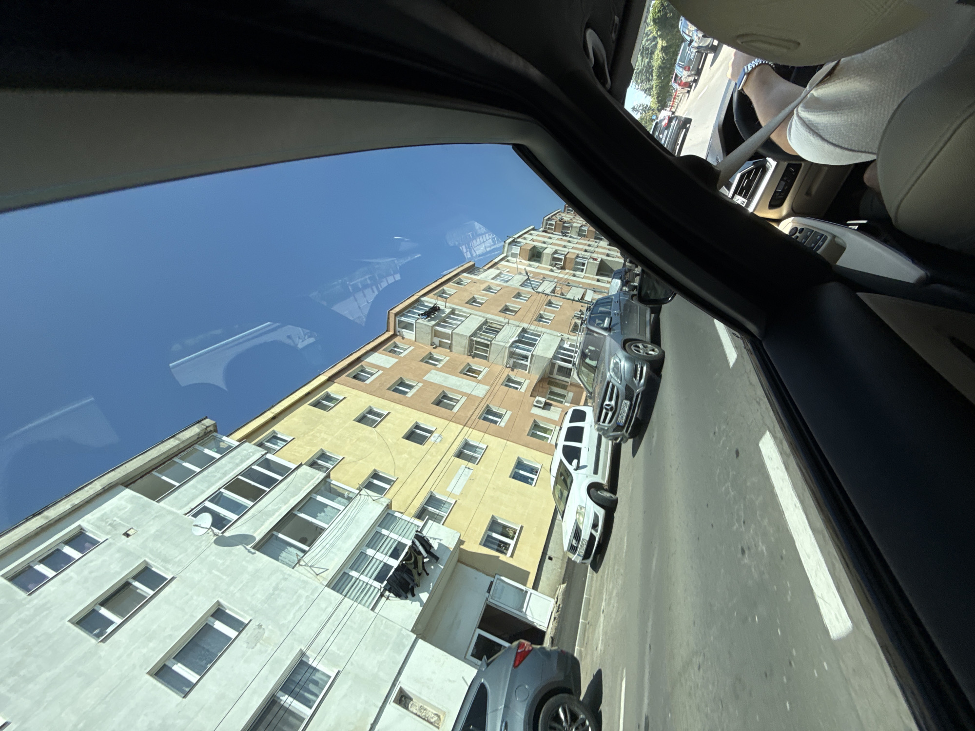 The image was taken from inside a vehicle, looking out through the sunroof and side window. The scene depicts a street in Târgu Secuiesc, Romania, with several apartment buildings lining the road. The buildings are of various colors, including pale green and yellow, and have multiple windows and balconies. Laundry is visible hanging from a balcony on one of the buildings.
Several cars are on the street, some parked and some in motion. A gray SUV is parked on the left side of the road, and a white minivan and a silver Mercedes-Benz SUV are visible in the street. The road has white lane markings. The sky is clear and blue. The interior of the vehicle is visible in the foreground, including the dashboard, steering wheel, and the driver's hands wearing a watch.