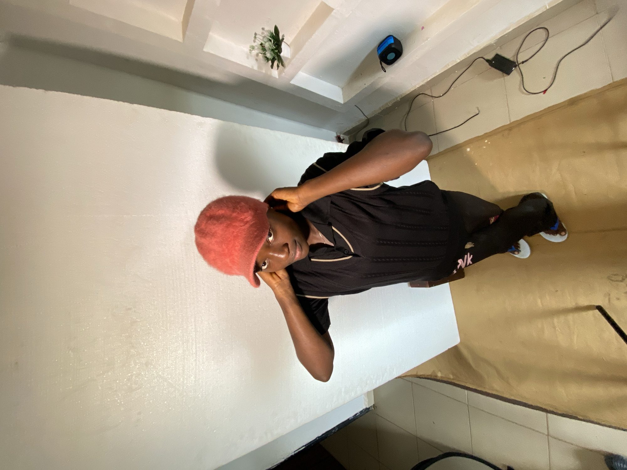 A young Black person wearing a black short-sleeved collared shirt and dark pants is posing with their hands on their head, wearing a fuzzy red cap. They are standing on a light brown floor in front of a white wall. To the right, there are white shelves built into the wall, one of which holds a small white pot with a green plant. Below the shelves, a small blue speaker and a black charger with a cord are visible on the floor. The scene is indoors, likely in a residential setting in Jos, Nigeria, as indicated by the tiled floor in the background and the general interior design.