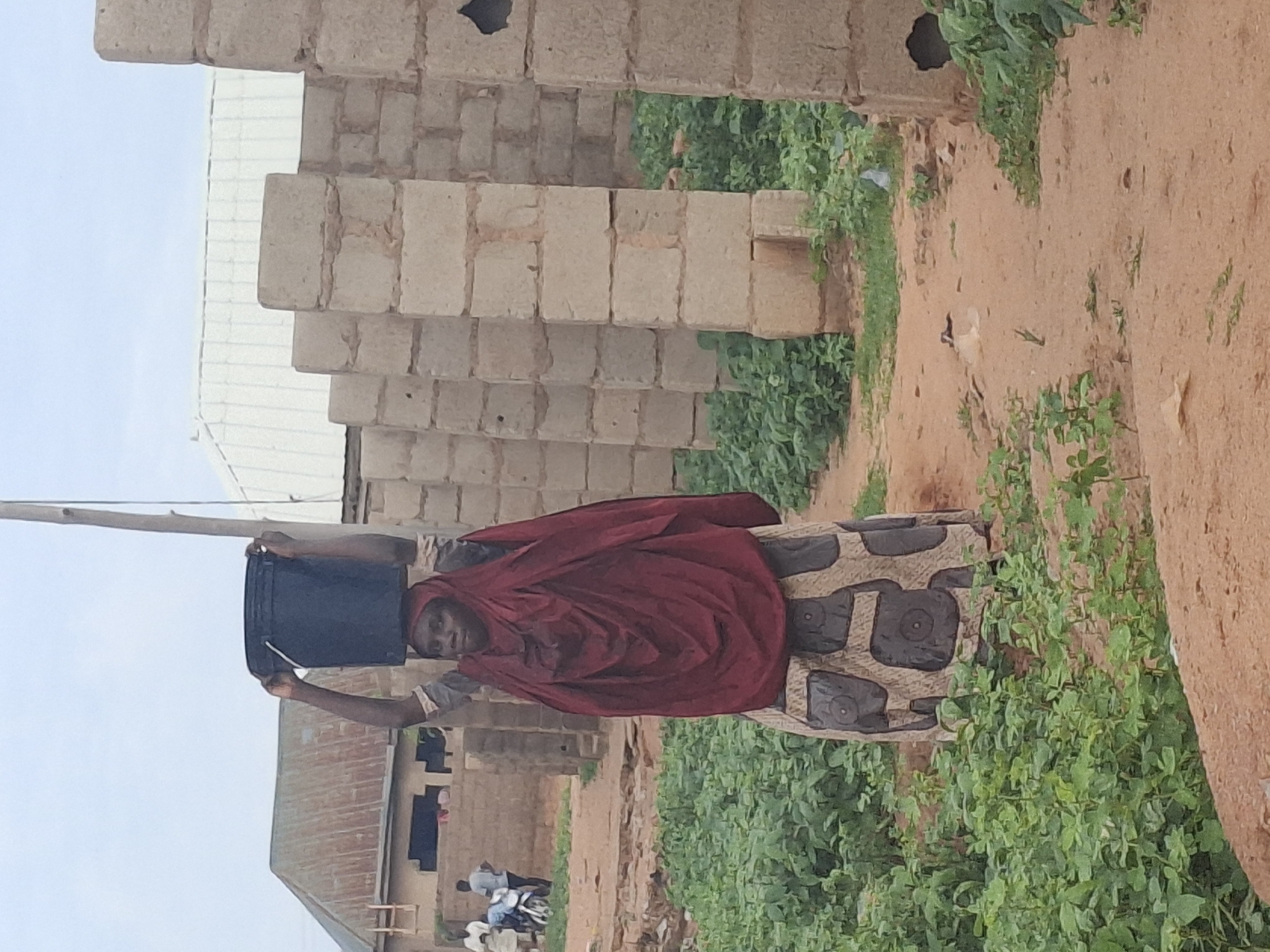 A woman in Tumfure, Nigeria, is captured in a full-body, vertical shot as she carries a black bucket on her head. She is wearing a maroon headscarf and a patterned dress with dark gray circular motifs on a lighter background. Her arms are raised to balance the bucket. The environment appears to be a construction site or an area with unfinished buildings, indicated by a row of concrete block pillars and walls. Green vegetation grows around the path. In the background, a building with a corrugated metal roof is visible, and further back, a few people and a motorbike can be seen. The ground is sandy, with some scattered debris. The sky is overcast.