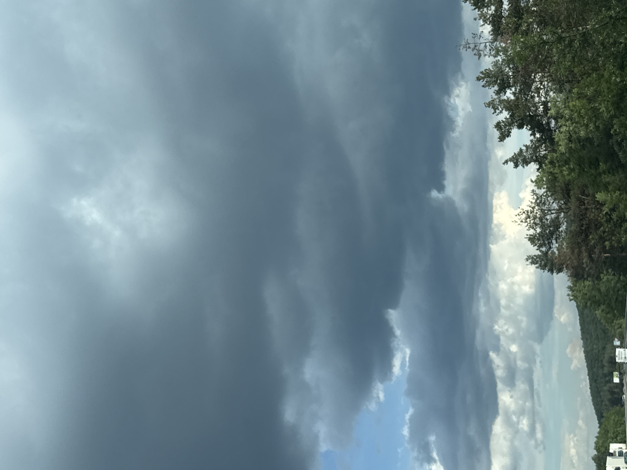 The image shows a view of the sky from a highway, with dark storm clouds dominating the upper portion. Below the clouds, there are patches of blue sky and lighter, more scattered clouds. The horizon is lined with green trees and a rolling hillside. On the highway, several white trucks and cars are visible, along with some road signs. The overall impression is of an overcast or approaching storm.