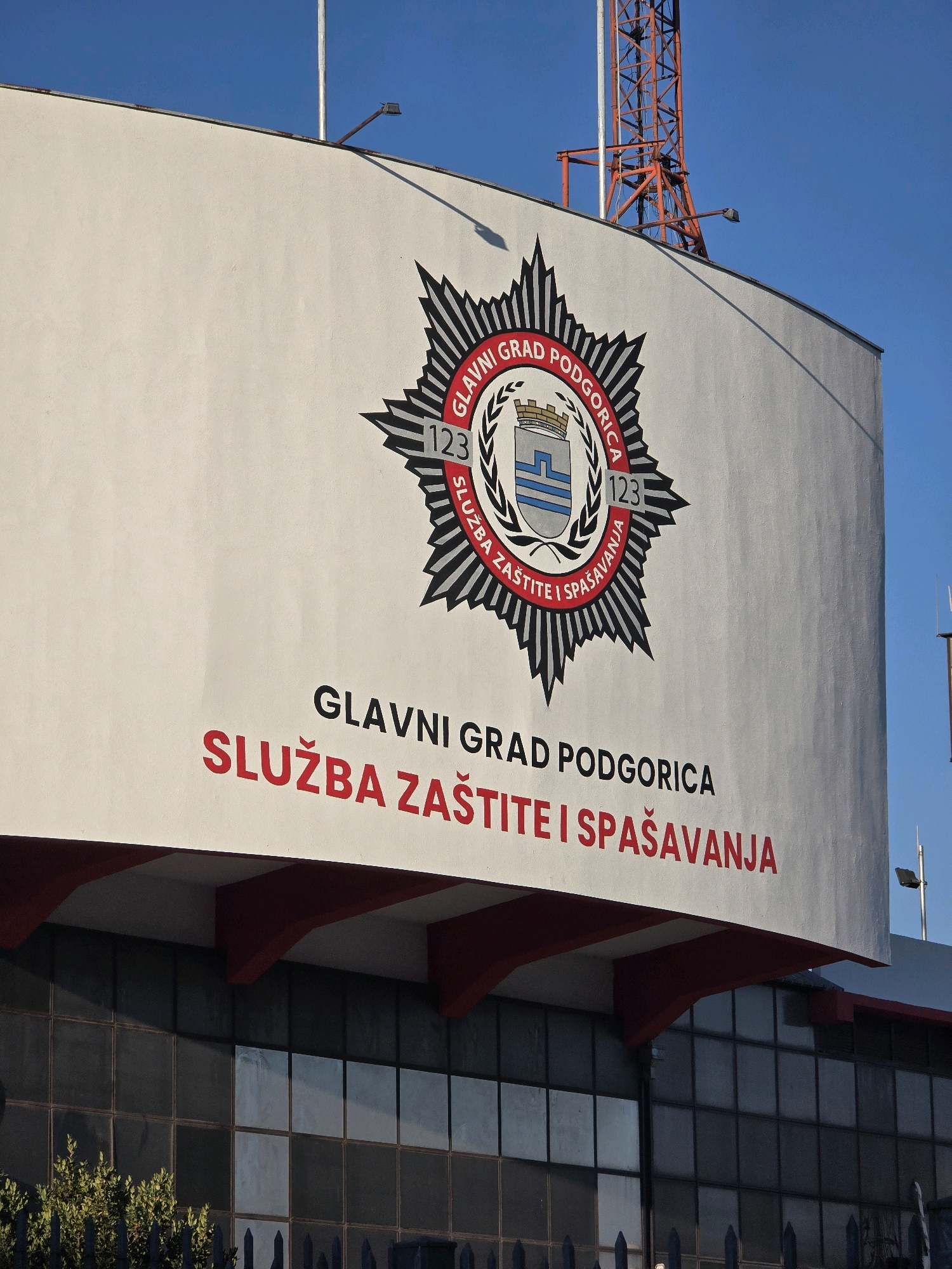 The image shows the exterior of a building with a large emblem and text painted on its facade. The emblem is a stylized star with various symbols and text arranged around it. Below the emblem, the text "GLAVNI GRAD PODGORICA" is written in black, followed by "SLUŽBA ZAŠTITE I SPAŠAVANJA" in red. This indicates the building is associated with the Protection and Rescue Service of Podgorica, the capital of Montenegro. The building appears to be modern, with large glass windows on the lower level. In the background, a radio tower and a clear blue sky are visible. The overall scene suggests an official government or public service building.