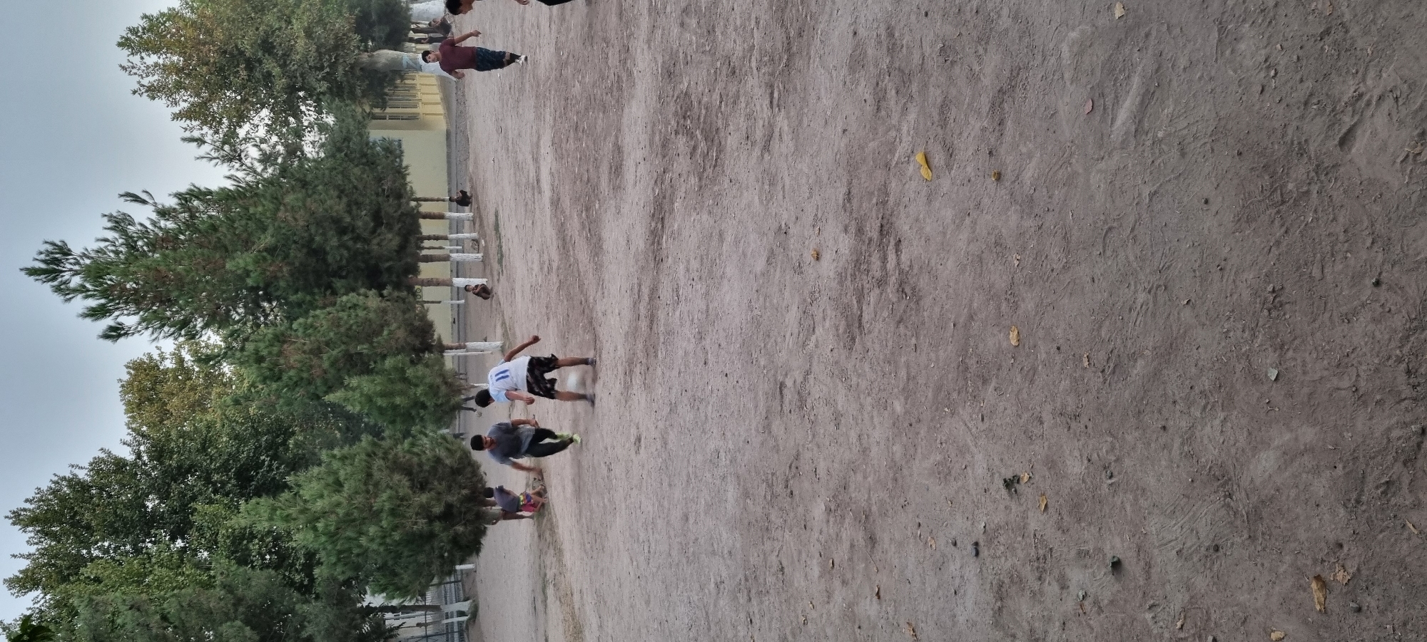 A group of young boys are playing soccer in a dusty, outdoor area in Хаваскор, Tajikistan. The field is largely unpaved, with scattered patches of dry grass and leaves. Several trees line the background, with a light-colored building visible behind them. The sky is overcast. In the foreground, two boys are actively engaged in the game, one in a white shirt with the number 11 on the back, the other in a gray shirt. Both are running and appear to be focused on the soccer ball. Other boys are scattered around the area, some observing and others possibly waiting their turn. One boy in a dark red shirt is walking in the middle ground. The overall scene depicts a casual recreational activity in a park or schoolyard setting.