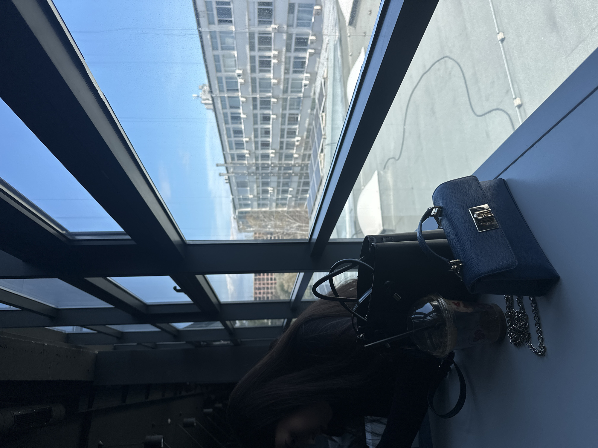 The image shows an interior scene with a partial view of a person with long, dark hair. A black purse and a blue, small handbag are placed on a light-colored surface, near a plastic cup with a straw, possibly containing a beverage. The background features a modern architectural structure with a glass ceiling and a building visible through the glass, suggesting an urban environment. Based on the architectural style, the location is likely in Sofia, Bulgaria.