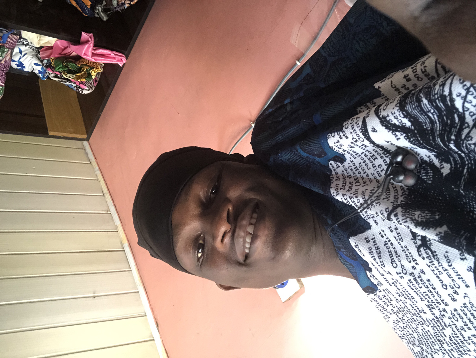 A young Black man wearing a black skullcap and a dark blue and white patterned shirt is taking a selfie. He is smiling and looking at the camera. The background shows a light-colored, paneled wall and a peach-colored floor. To the right, a shelf is filled with colorful fabrics. The setting appears to be indoors, possibly a home or a shop. The man is in the city of Ado, Nigeria.