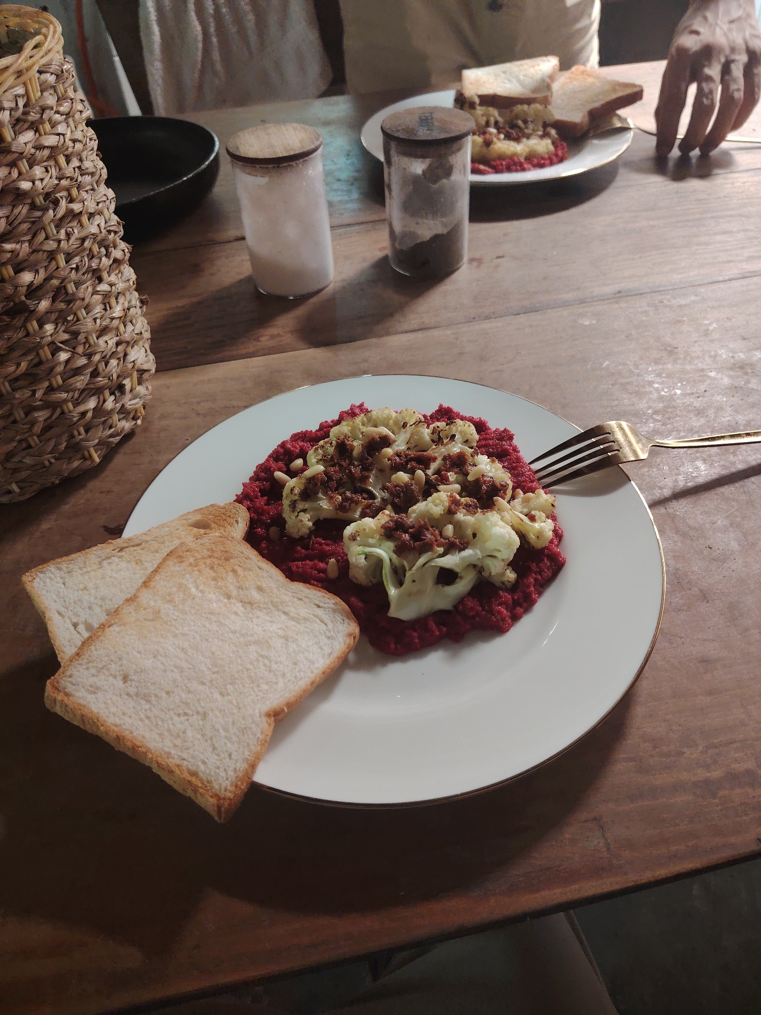 An overhead shot captures a cozy meal laid out on a rustic wooden table. In the foreground, a white plate with a gold rim holds a vibrant red purée, likely beetroot, topped with roasted cauliflower florets, scattered pine nuts, and what appears to be sundried tomato bits or a dark spice mixture. Two slices of lightly toasted white bread rest against the plate, ready to be enjoyed with the colorful dish. A gold-toned fork lies on the right side of the plate.
In the background, another similar meal is visible on a white plate, also with toast, suggesting two people are dining. A person's hand is visible on the right, near the second plate, as if reaching for food. The upper body of a person, possibly wearing a light-colored shirt, is partially visible behind the second plate. A dark, possibly empty, bowl and two clear glass jars with wooden lids are scattered on the table; one jar contains a light-colored substance like salt, and the other holds a darker, finely ground spice like pepper.
To the left, a tall, woven straw basket adds a natural texture to the scene. The wooden table itself shows grain and some wear, contributing to the rustic ambiance. The overall lighting is dim and warm, indicating an evening setting, creating an intimate and comfortable atmosphere for the meal. No visible text is present in the image.
