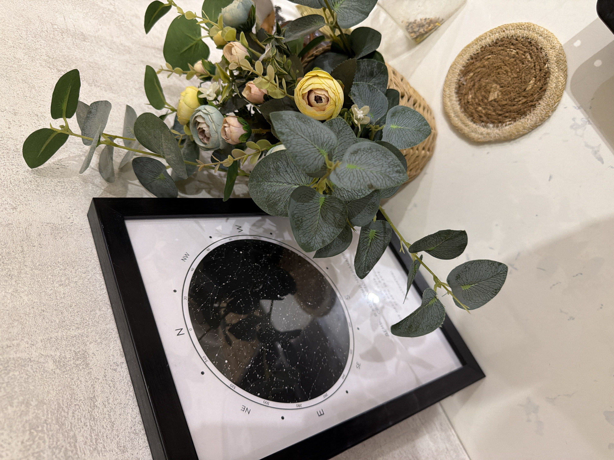 The image displays a framed star map and a small basket of artificial flowers, placed on a light-colored, speckled surface, possibly a countertop. The star map, enclosed in a black frame, features a dark background with white dots representing stars, and a compass rose with directional labels such as N, NW, W, SW, S, SE, E, and NE. The background wall has a textured, light beige appearance. To the right of the framed map, the basket contains a floral arrangement with various artificial flowers in shades of pink, yellow, and light blue, interspersed with green eucalyptus leaves. A woven coaster with a spiral pattern sits on the surface to the right of the flower basket. A small, clear plastic bag containing what appears to be seeds or small grains is also visible. The scene appears to be indoors, likely in a domestic setting, with soft lighting casting subtle shadows. There is no indication of the city of Semiluki, Russia, or any specific geographical location.