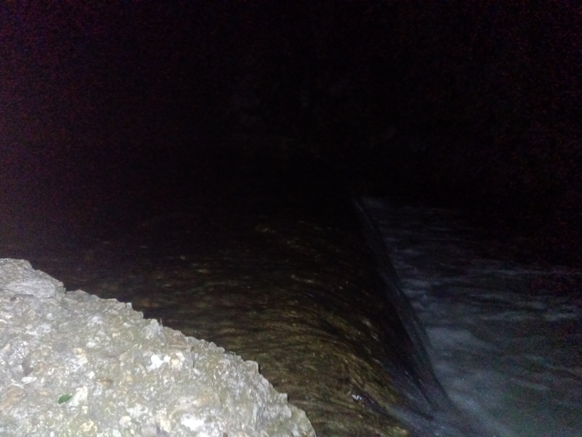 This media file presents a dark, low-light scene, likely captured at night, depicting a body of water flowing next to a rocky bank. In the bottom left, a rugged, light-colored bank made of rocks or concrete is prominent, displaying varied textures and shades of gray and white, with a small green leaf visible among the rocks. The water, which appears dark, occupies the central and right portions of the image. Subtle patterns on the water's surface suggest movement and current. Towards the right, the water flows over a sharp, linear edge, creating a small waterfall or weir, where the water appears frothy and lighter in color as it cascades downwards. The upper parts of the image are extremely dark, providing minimal detail and emphasizing the low-light conditions. No people or visible text are present. The setting is an outdoor environment in Tahlequah, United States of America, likely featuring a river, stream, or other water channel.