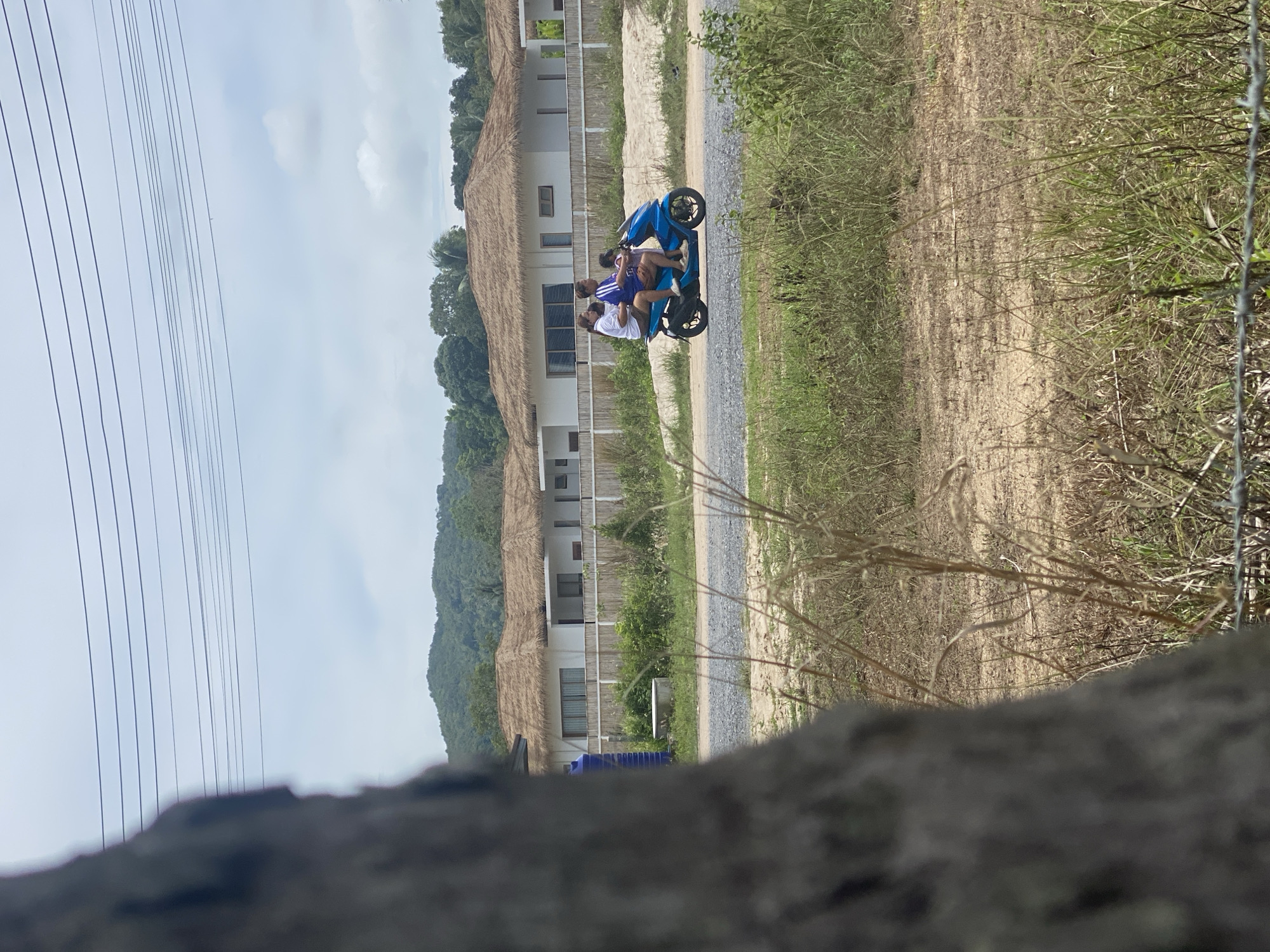 In Moo 1, Thailand, the image captures a scene with two individuals on a blue scooter traversing a gravel path. The scooter is the primary subject, and the two riders are positioned on it. To the right of the path is vegetation, and behind the scooter are buildings, possibly resorts or accommodations, with thatched roofs. In the background, a hill or mountain is visible. The foreground shows a portion of a wall or stone structure, partially obscuring the view. Power lines stretch across the upper-left portion of the frame.