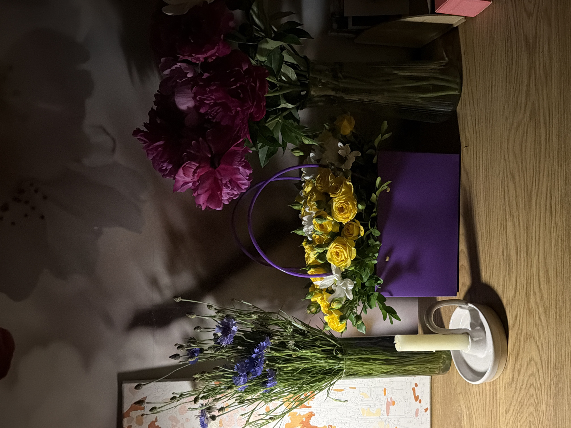 The image depicts a tabletop still life. The table, made of light-colored wood, is oriented vertically in the frame. A bouquet of dark pink peonies dominates the upper-left portion of the scene. Another bouquet, composed of yellow roses and small white flowers, is partially contained within a purple gift bag. Below these, a bouquet of blue cornflowers lies near a small canvas or board with abstract orange and white patterns. A white ceramic candle holder with a cylindrical candle sits near the right edge of the scene. The location and city are unknown.