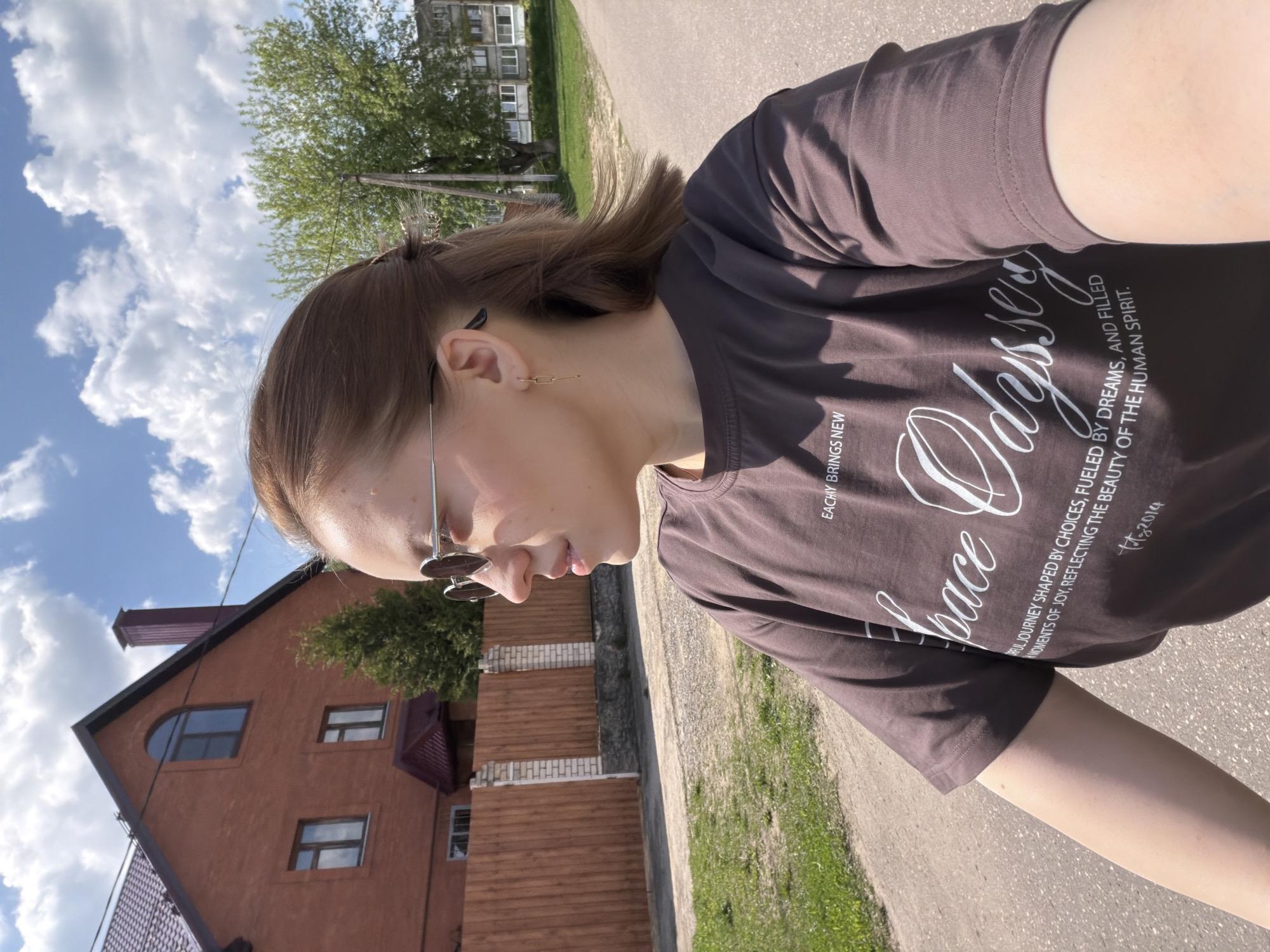 The image shows a woman in Reshetnikovo, Russia, standing outdoors on a paved road. She is wearing sunglasses and a brown t-shirt with the words "Odyssey" and "EACHAY BRINGS NEW" printed on it. The shirt also features the phrase "A FUL JOURNEY SHAPED BY CHOICES, FUELED BY DREAMS, AND FILLED MOMENTS OF JOY, REFLECTING THE BEAUTY OF THE HUMAN SPIRIT." Behind her is a reddish-brown brick building and a wooden fence. A grassy area and trees are visible to the side of the road. The sky is visible in the upper left with clouds.