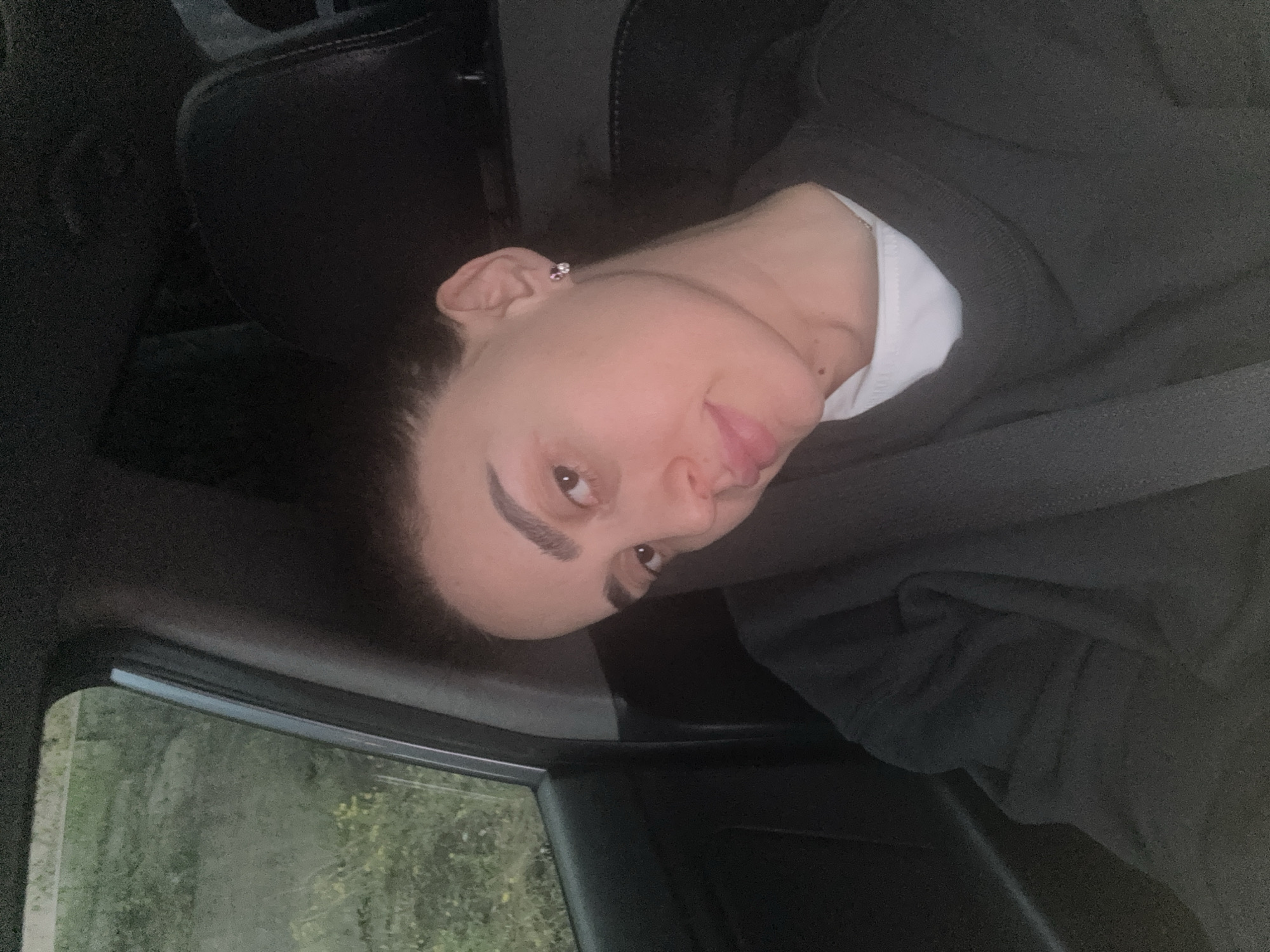 A person is sitting in the driver's seat of a car. The person has light skin, dark hair pulled back, and wears a seatbelt, a dark sweatshirt, and a white t-shirt visible at the neckline. The car is moving, as suggested by the blurred greenery visible through the open window. The location is unknown, but the prompt suggests it is Tbilisi, Georgia.