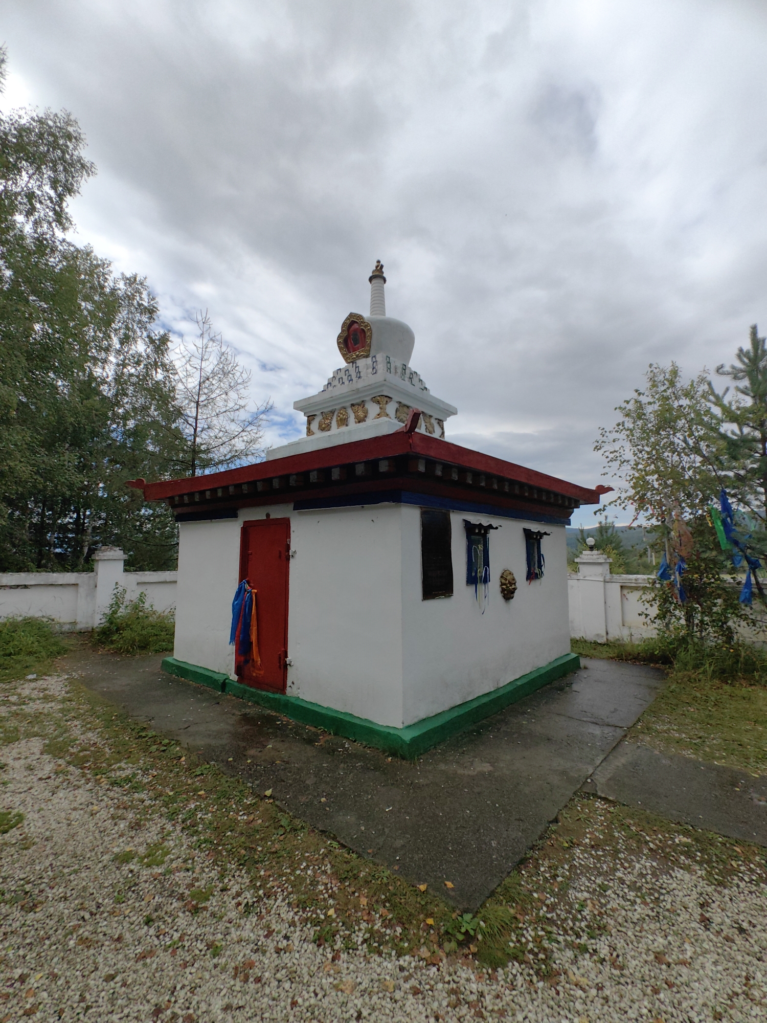 A white, rectangular Buddhist stupa stands in a courtyard with a gravel groundcover and some sparse vegetation. The stupa has a red roof with blue trim, a green base, and a white, tiered spire topped with a golden ornament. A red door is visible on the front of the stupa, with blue and orange prayer flags hanging from it. Two windows with blue frames are on the side of the building. The stupa is fenced by a low, white wall. Trees are visible in the background. The sky is cloudy. This structure appears to be located in a rural or semi-rural area, possibly part of a monastery or a religious site in the city of Tibelti, Russia.