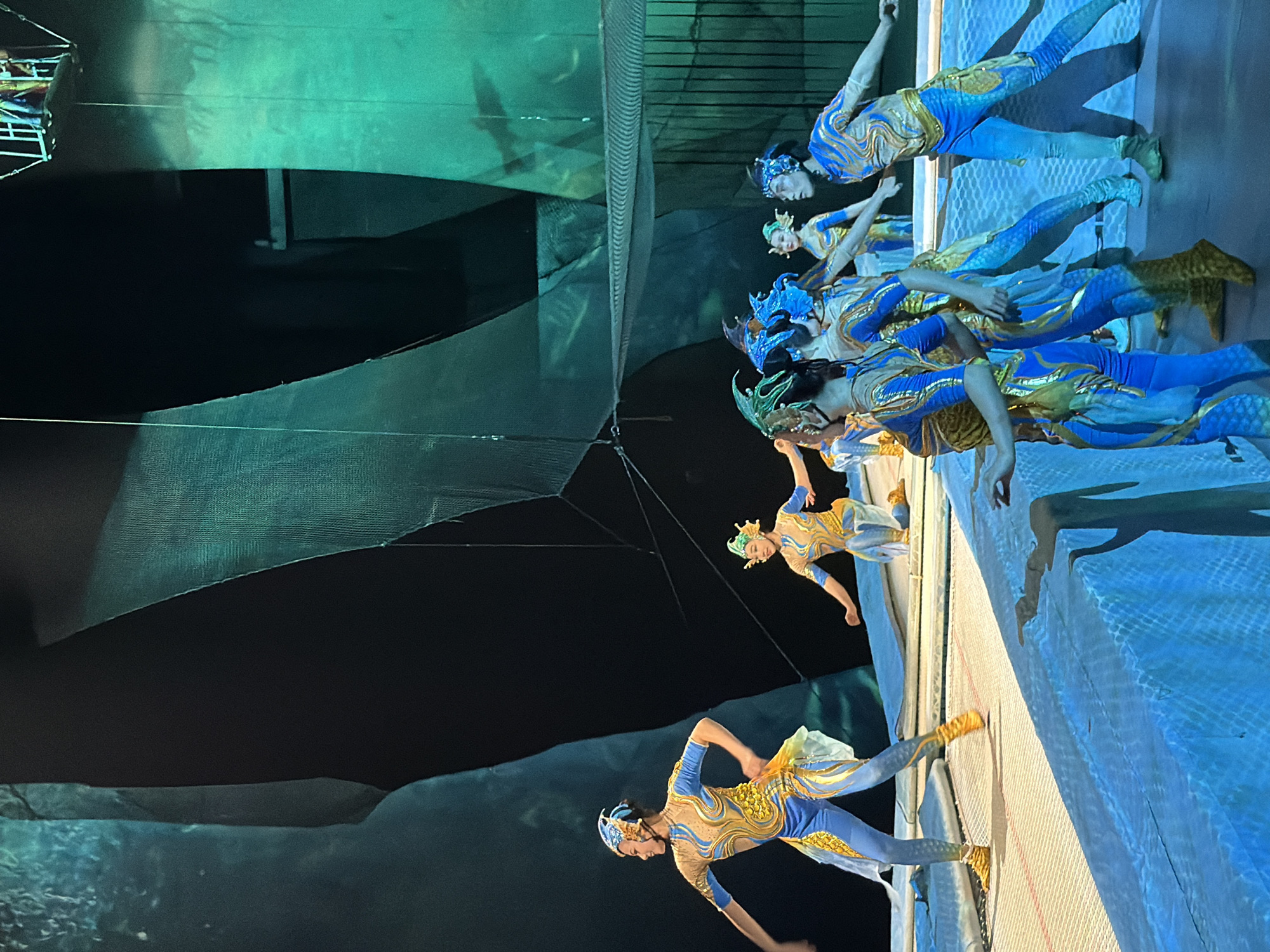 The image depicts a performance on a stage, likely within the city of Nan'an District, China. A group of performers, appearing to be young women and a young man, are dressed in vibrant blue and gold costumes that resemble aquatic creatures, possibly mermaids or mythical sea beings, complete with elaborate headpieces. They are positioned on a trampoline-like surface, with some in mid-action, suggesting a dynamic performance involving acrobatics or dance. The stage is lit with dramatic lighting, primarily blue and green, creating an atmospheric effect. The background features dark, abstract shapes and a large netted structure suspended from above, contributing to a theatrical and immersive environment.