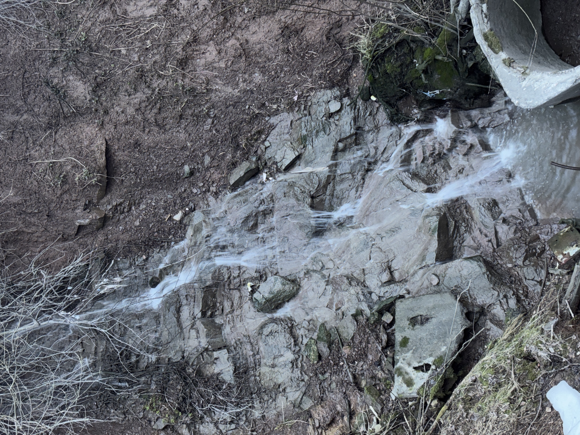 The image depicts a small waterfall cascading over rocks and debris. The setting is a natural landscape with exposed earth and sparse vegetation. A section of concrete piping is visible on the upper right of the image. The water appears cloudy or silty. The location is the urban area in Perm, Russia, which is the source of the small river that feeds the waterfall.
