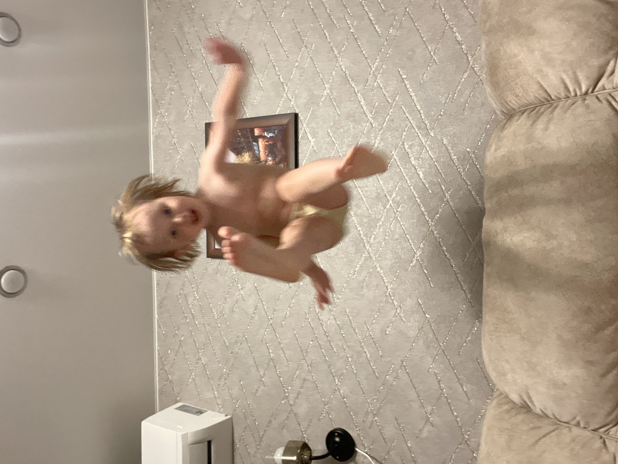 The image shows a young child, appearing to be a toddler, in mid-air, seemingly jumping or being thrown upwards. The child has light-colored hair and is wearing a light-colored diaper. Their arms and legs are outstretched in a dynamic pose, suggesting movement and energy. The child's face is turned towards the camera, with a slight smile or surprised expression.
The background features textured wallpaper with a subtle diamond pattern and some glittery elements. A framed picture hangs on the wall behind the child, though its contents are indistinct. To the right of the frame, a portion of a large, light-brown upholstered piece of furniture, likely a sofa or armchair, is visible. On the left side of the image, a white wall with a circular fixture is present. In the lower part of the image, there's a white rectangular object, possibly an air purifier or humidifier, and a small black object with a light bulb, which could be a lamp.
The lighting suggests it is likely daytime indoors, as there are no strong shadows to indicate evening or night. The overall mood appears playful and energetic, capturing a moment of childhood exuberance. No visible text is present in the image. The location context of Vladivostok, Russia, is provided but not discernible from the visual elements of the image itself.