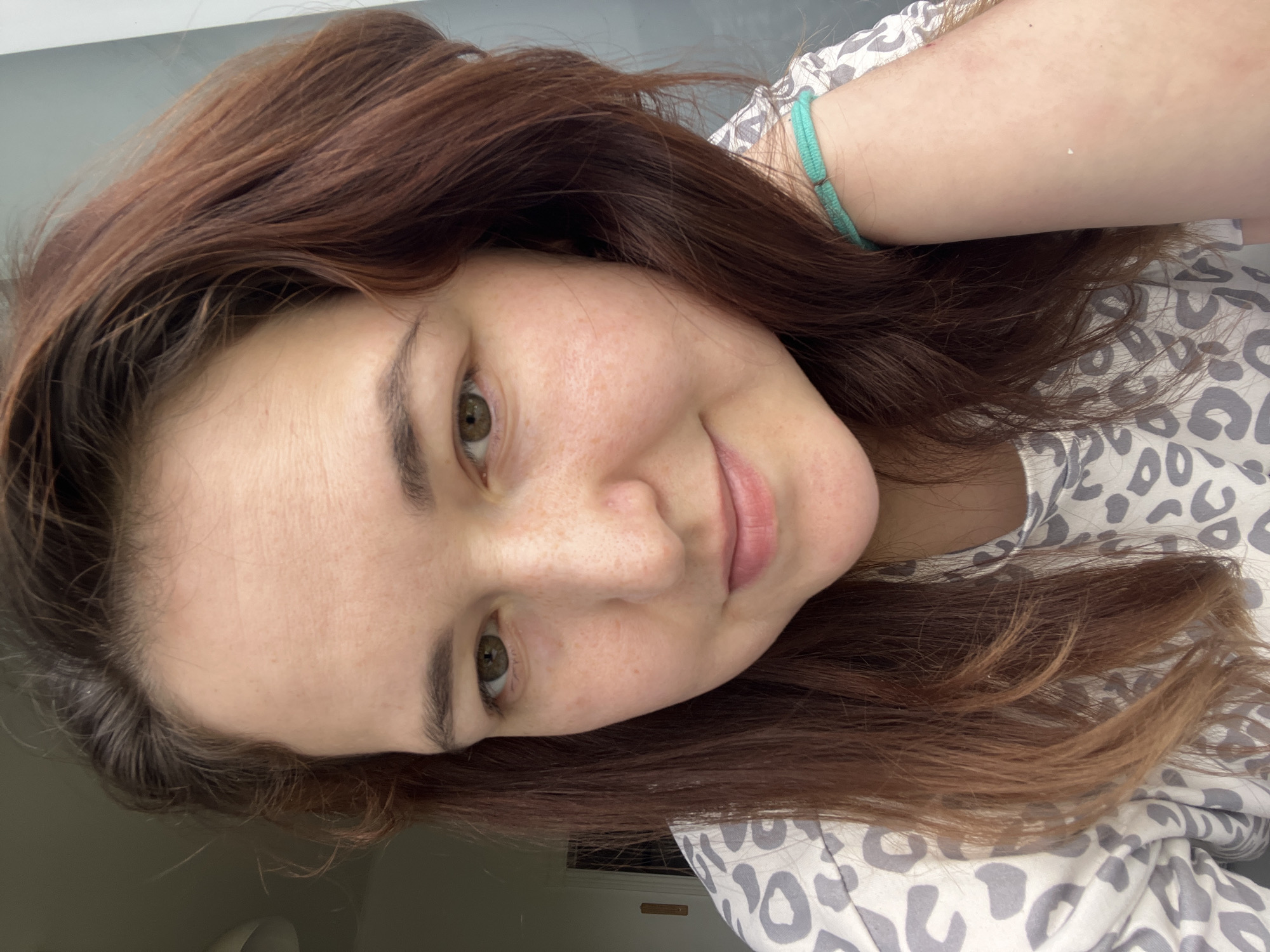 The media file is a close-up, self-taken photograph of a young woman with long, auburn hair. She is looking at the camera with a slight smile. Her eyes are a green-brown hue, and she has light skin with a scattering of freckles. She is wearing a light-colored shirt with a grey and white leopard-like print. A thin, teal bracelet is visible on her left wrist. The lighting suggests it is daytime, and the indoor setting appears to be domestic. The woman's expression conveys a relaxed and pleasant mood. There is no visible text in the image.