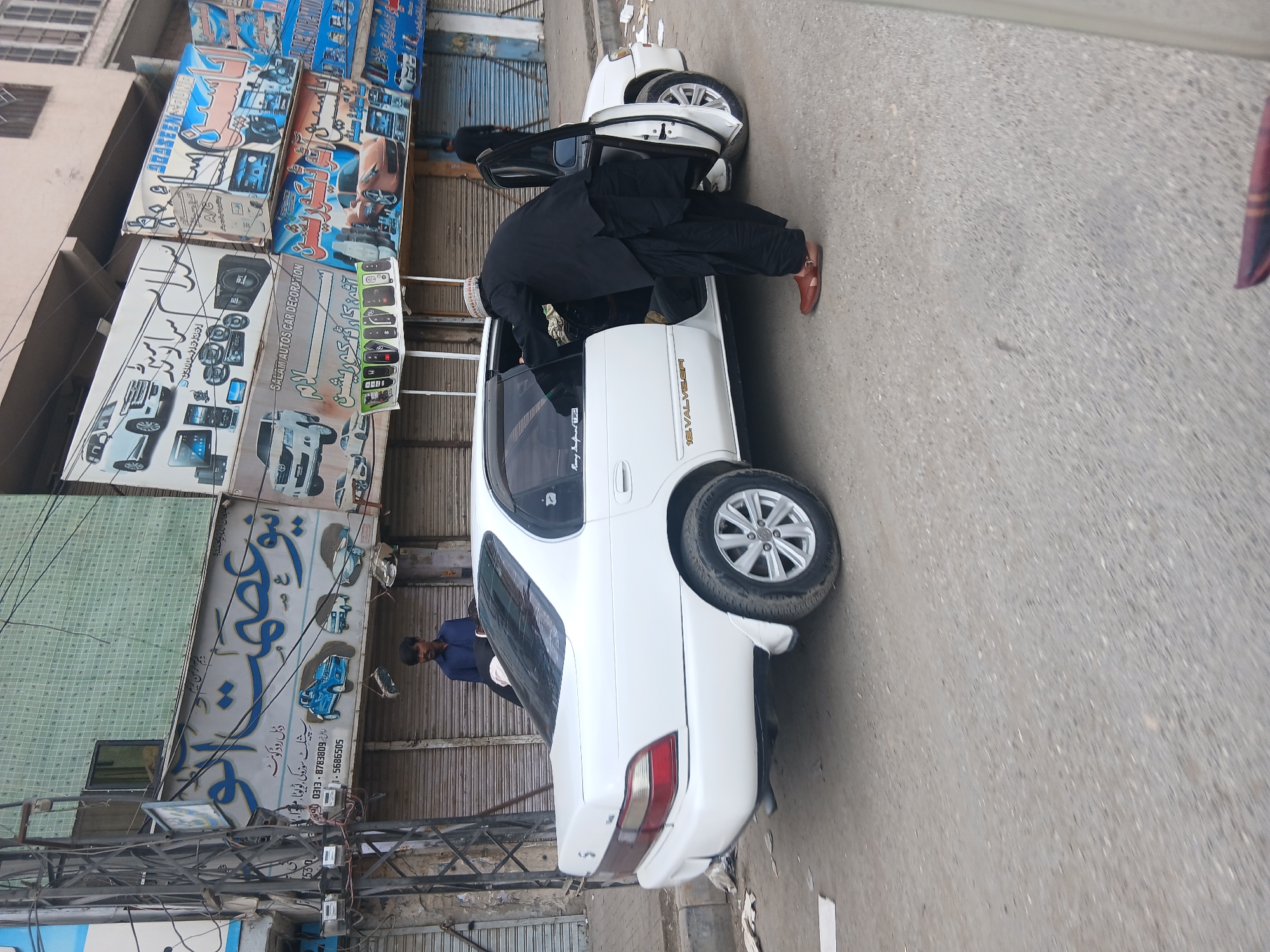 A man in a black shalwar kameez is bending into a white Toyota Corolla sedan parked on a street in Quetta City Tehsil, Pakistan. The car's passenger-side door is open. Another man in a blue shirt stands behind the car. Behind the car are shopfronts with corrugated metal doors and signs in Urdu for car parts and repair shops. Power lines and utility poles are visible. The street is paved and dry.