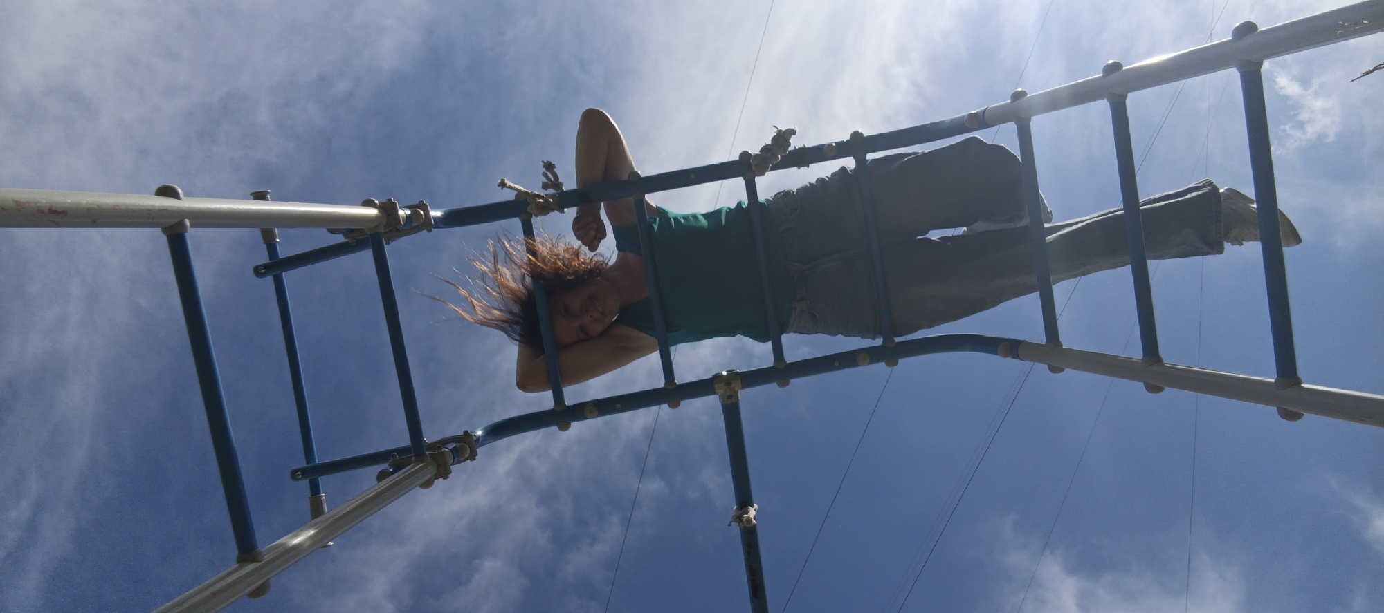 A low-angle shot captures a person, dressed in a teal shirt and grey pants, hanging upside down from a monkey bar structure against a bright blue sky with scattered white clouds. The person's hair is a reddish-brown, and their face is turned slightly towards the camera, with eyes open and a slight smile. The monkey bars appear to be made of metal and are part of a playground. The overall scene is outdoors, with the sky dominating the background. The location is specified as Chaikovsky, Russia.