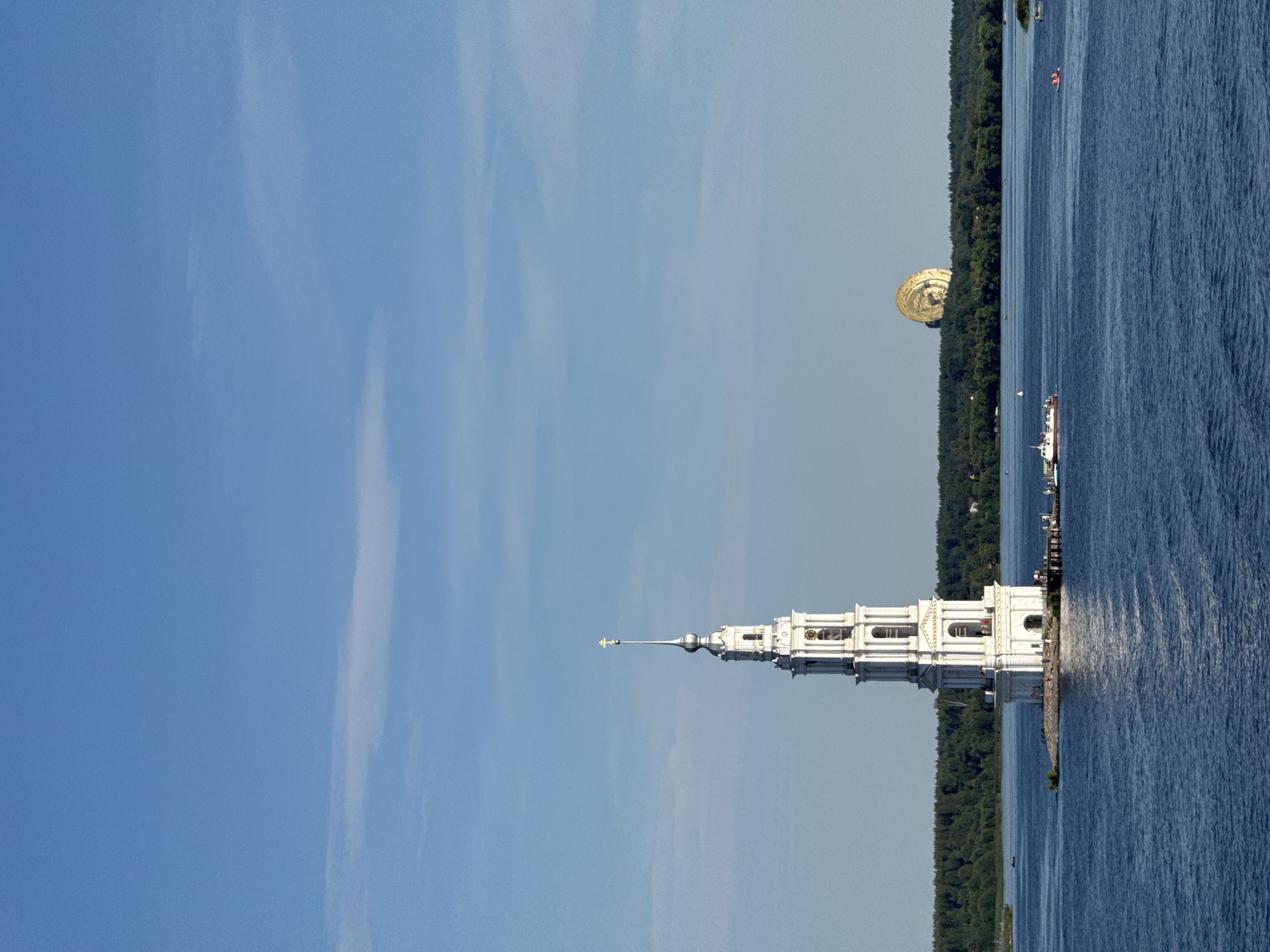 A white, multi-tiered bell tower stands on a small island in the middle of a wide, blue body of water. The tower has a tall, slender spire topped with a golden cross. The lower portion of the tower is wider and rectangular, with arched windows and entrances. People are visible on the island and a pier extending from it. A few small boats are on the water, including a white boat with people on board. In the background, a dense green forest lines the far shore. Further in the distance, a large, golden, disc-shaped structure with a patterned surface is visible above the treeline. The sky is a clear, pale blue with a few wispy clouds. The bell tower is the Kalyazin Bell Tower, an iconic landmark in the city of Kalyazin, Russia, submerged by the creation of the Uglich Reservoir in 1939.