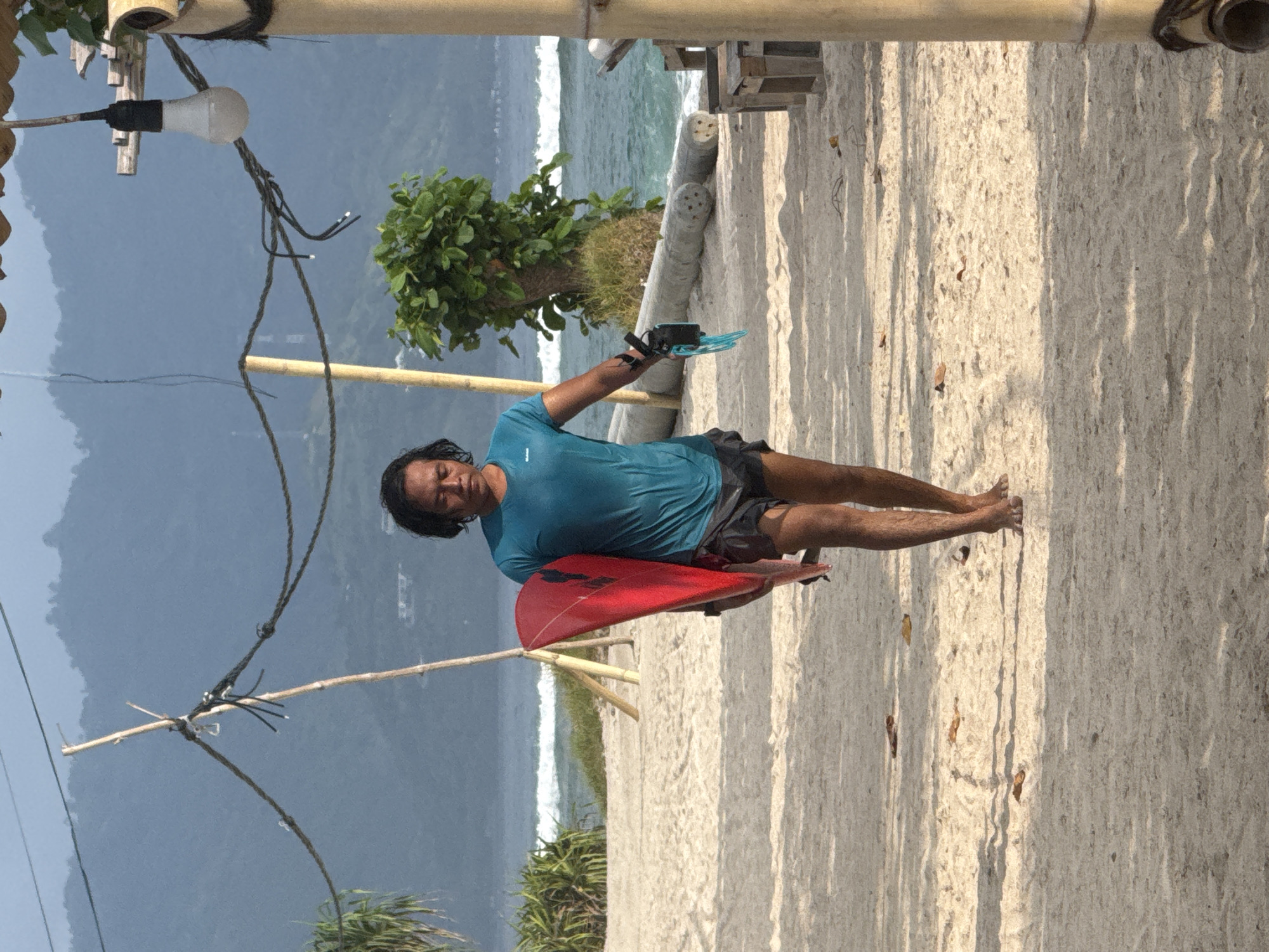 The image captures a man standing on a sandy beach, holding a red surfboard under his left arm. He wears a teal or light blue short-sleeved t-shirt and dark shorts. His dark, medium-length hair frames his face as he looks towards the viewer with a neutral expression.
The setting appears to be a sunny day at a tropical beach. Strong shadows are cast on the light-colored sand, indicating bright sunlight. To the man's right, the ocean stretches out with white-capped waves visible breaking towards the shore. Further in the background, lush green foliage and bushes line the coastline, and large mountains rise under a clear, pale blue sky.
Above the man's head, part of a rustic structure made of bamboo poles is visible. A simple light bulb hangs from a wire attached to this structure, along with other electrical wiring or ropes. Another thin bamboo pole crosses horizontally in the mid-ground. The overall scene suggests a vibrant beach environment, possibly a surf destination. There is no visible text in the image.