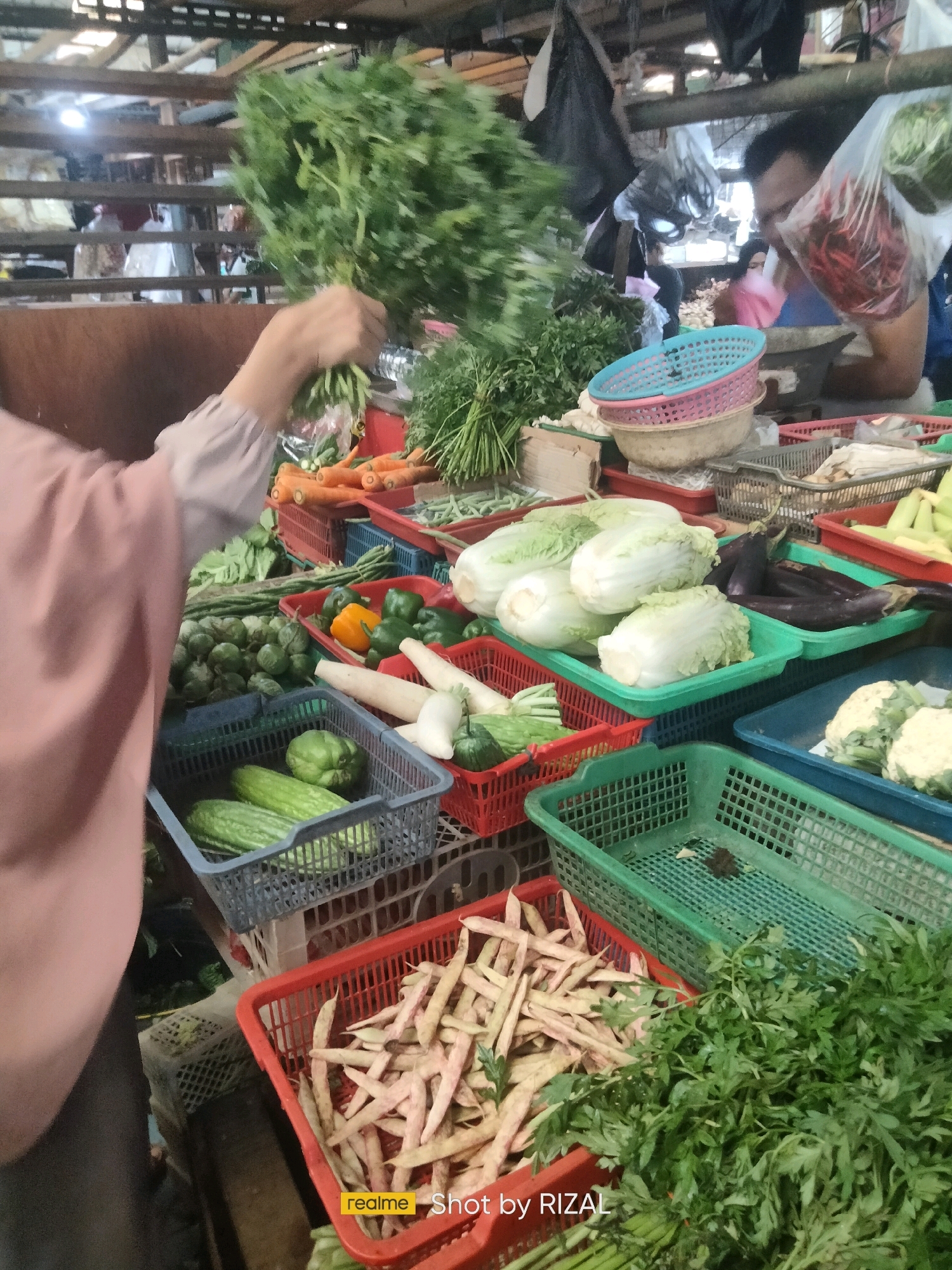 This image depicts an outdoor market scene in Bojong Nangka, Indonesia, likely a wet market. In the foreground, a person wearing a light pink top is holding a large bunch of leafy green vegetables. Various fresh produce is displayed in baskets and crates on a table. These include carrots, green and yellow bell peppers, cucumbers, leafy greens such as bok choy, and string beans. In the background, other market vendors and customers are visible, with stalls selling more produce, some in plastic bags. The environment appears to be covered by a wooden roof structure.