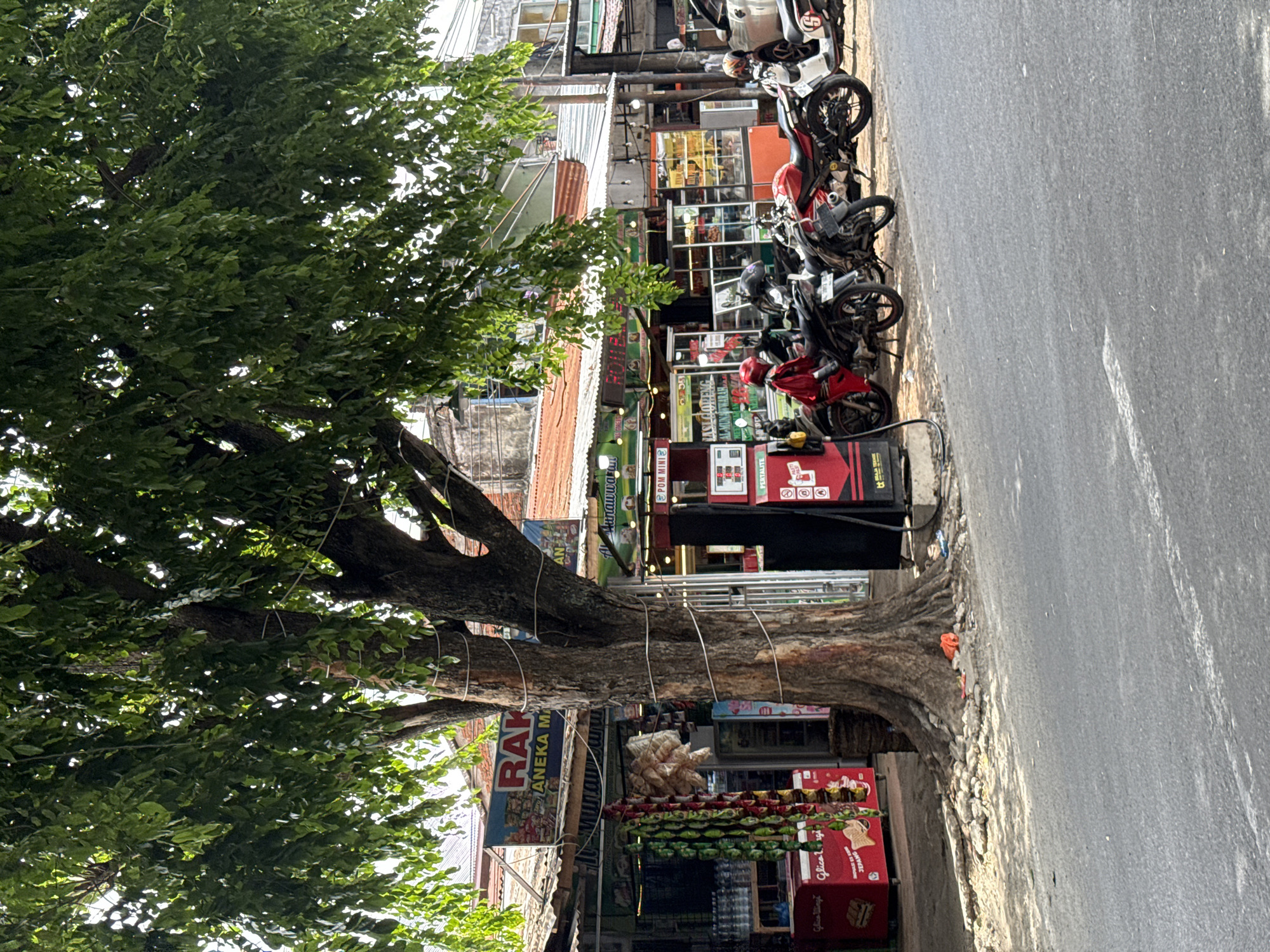 In Siron, Indonesia, a street scene unfolds under the shade of a large tree. Several motorcycles are parked along the roadside in front of a row of shops. One motorcycle, a red sportbike, has a rider wearing a red jacket and helmet. Another motorcycle, a white scooter, is parked further down. A red gasoline pump is positioned near the parked motorcycles. The shops behind the motorcycles appear to be selling various goods, with some displaying signs with Indonesian text. One sign reads "RAKA ANEKA MAKMUR." Bags of snacks hang from a display in front of one of the shops, and a red refrigerator with the brand "Glicoo" is visible. The environment is a bustling street with buildings, a tree, and parked vehicles, suggesting a commercial area.